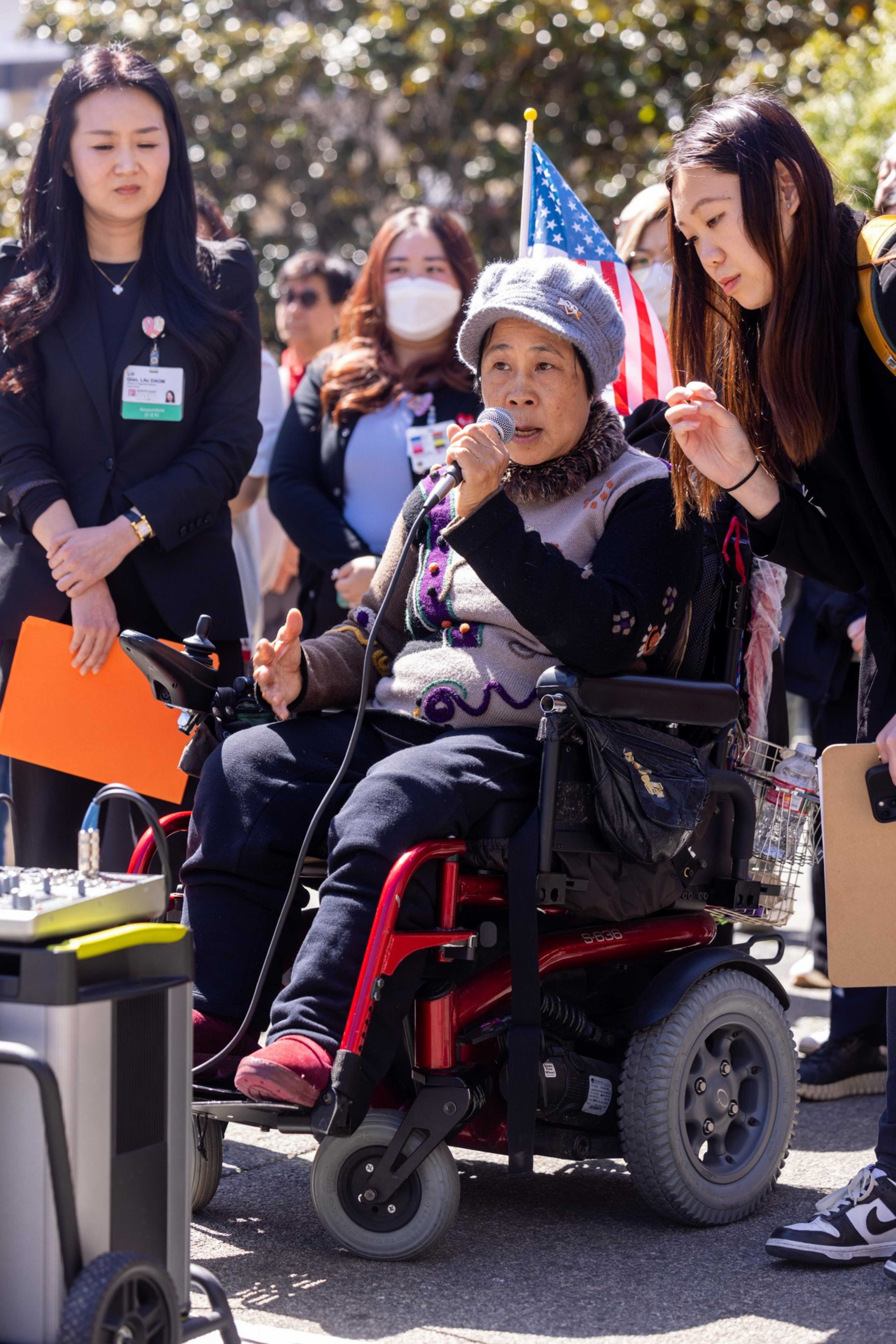 An elderly woman in a wheelchair speaks into a microphone, surrounded by women, one holding a clipboard. An American flag is attached to her chair.