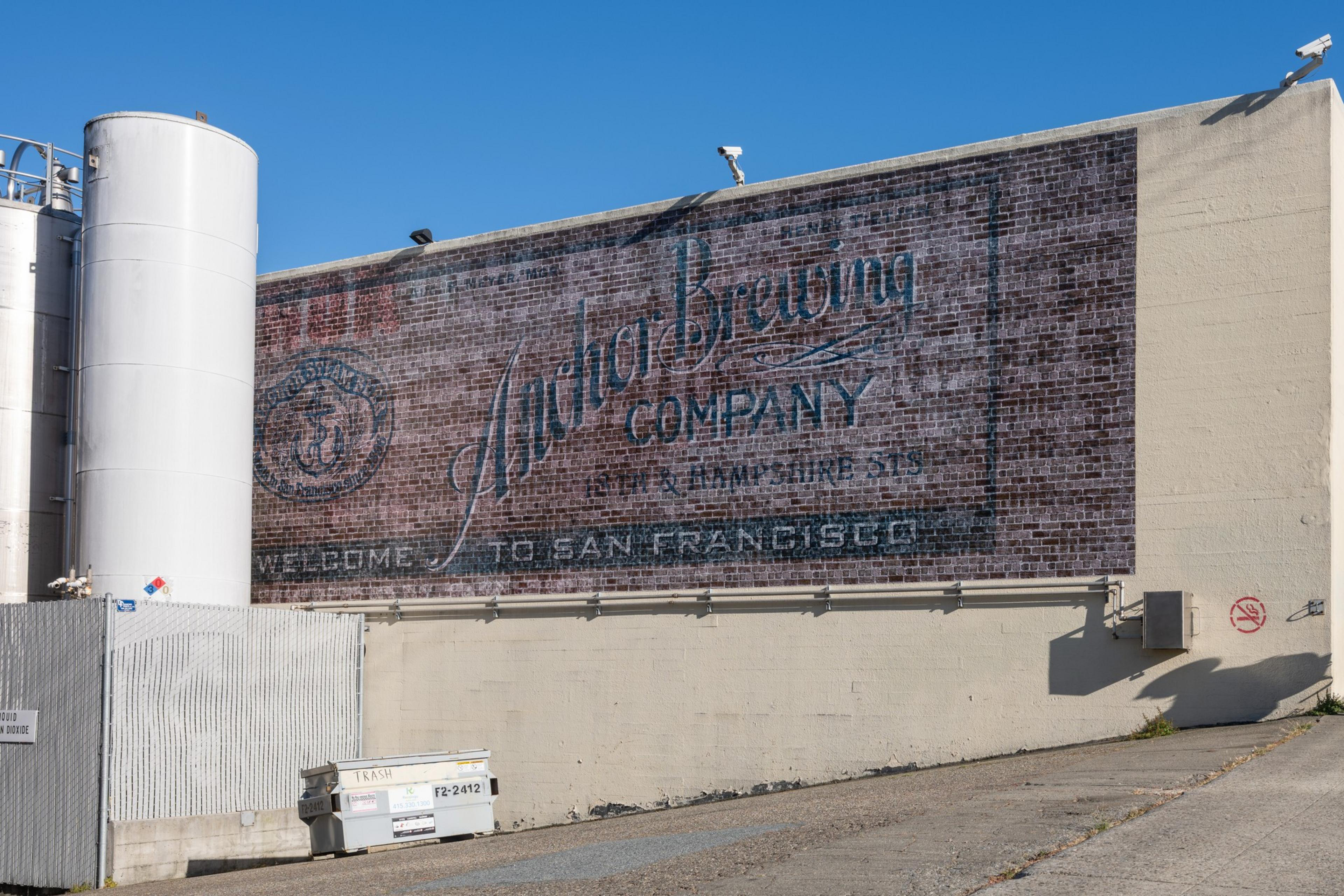 A faded mural on a brick wall reads &quot;Anchor Brewing Company&quot; with &quot;Welcome to San Francisco.&quot; Nearby are large white tanks and a small trash dumpster.