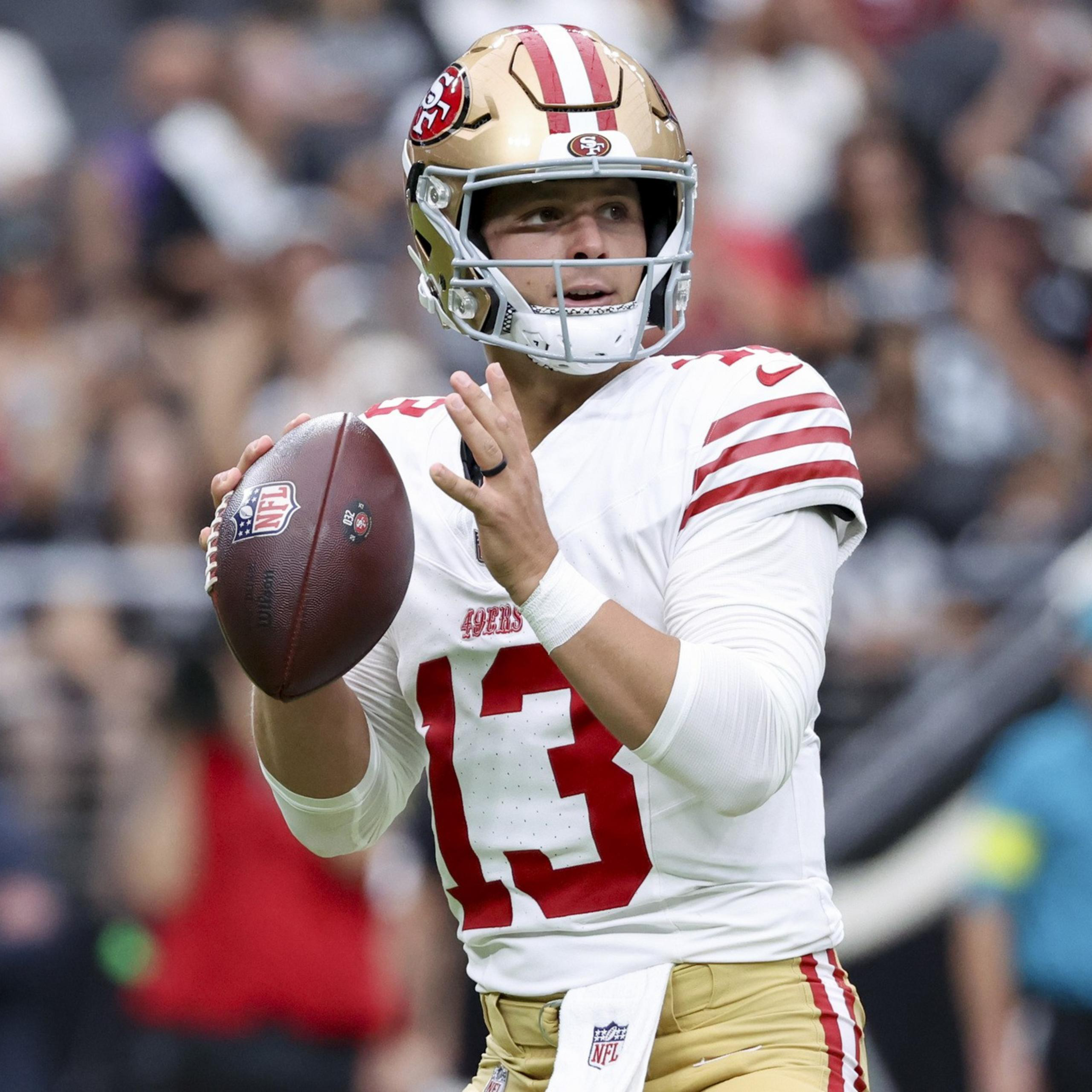 A football player in a white and gold 49ers uniform, number 13, prepares to throw a pass during a game with a blurred crowd in the background.