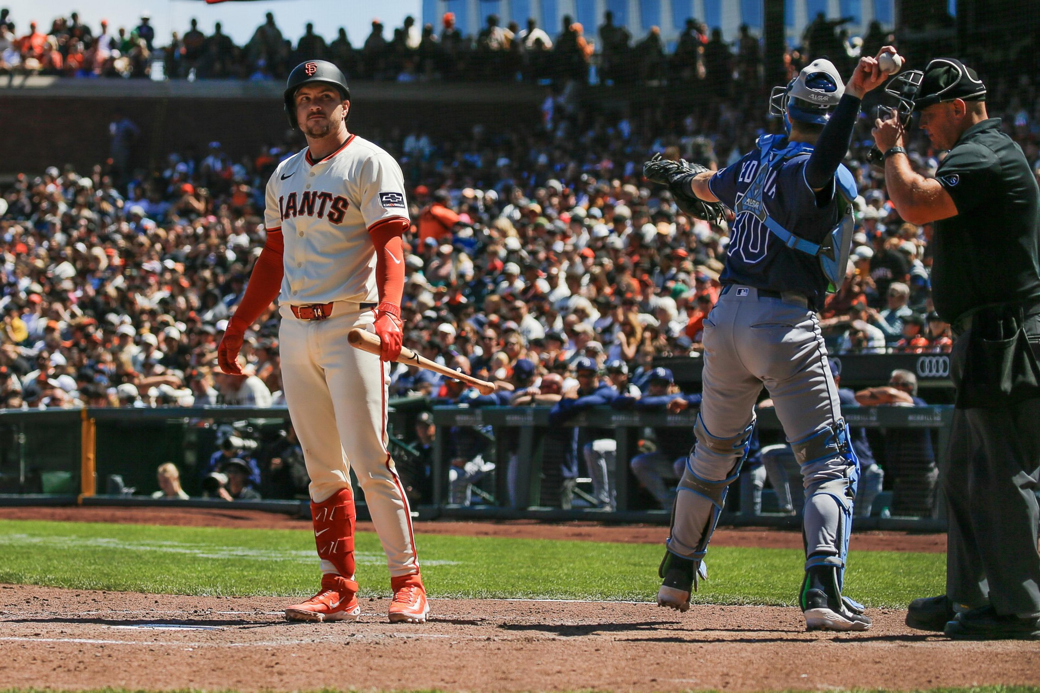 A San Francisco Giants batter stands at home plate holding a bat, while a catcher in gray and blue gear throws a pitch behind him as the umpire watches closely.