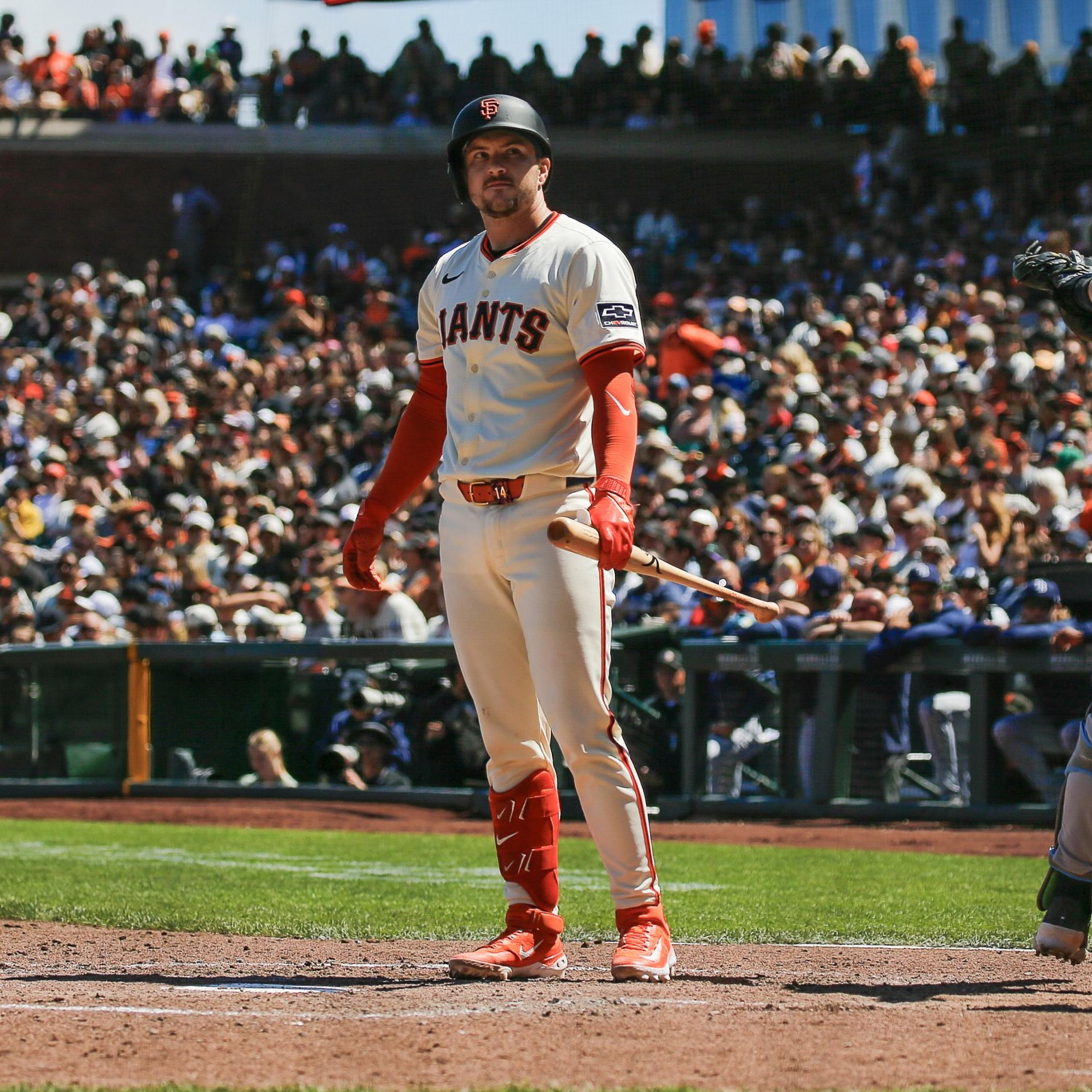 A San Francisco Giants batter stands at home plate holding a bat, while a catcher in gray and blue gear throws a pitch behind him as the umpire watches closely.