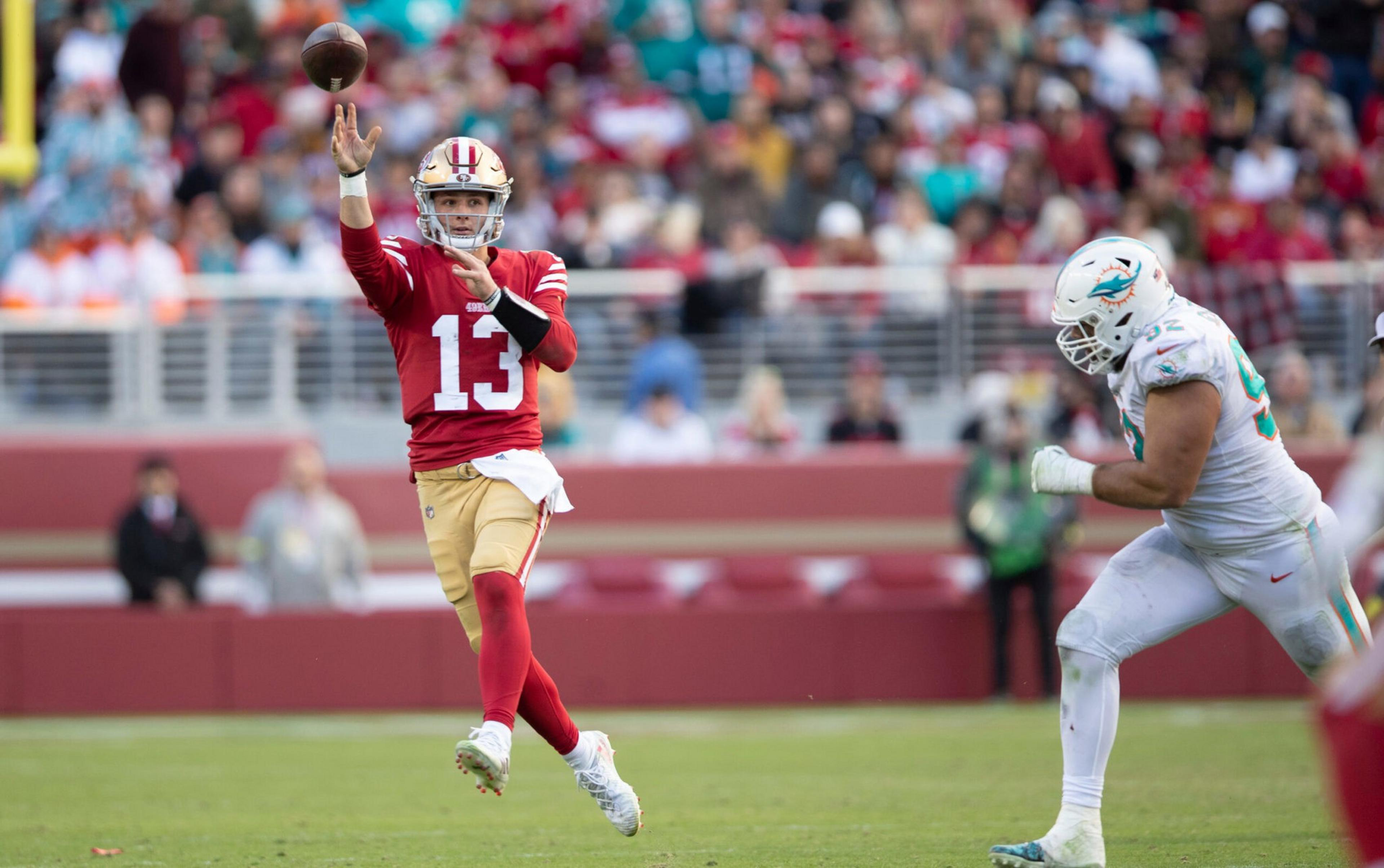 A football player in a red and gold uniform throws a pass while a player in a white uniform with a dolphin logo approaches.