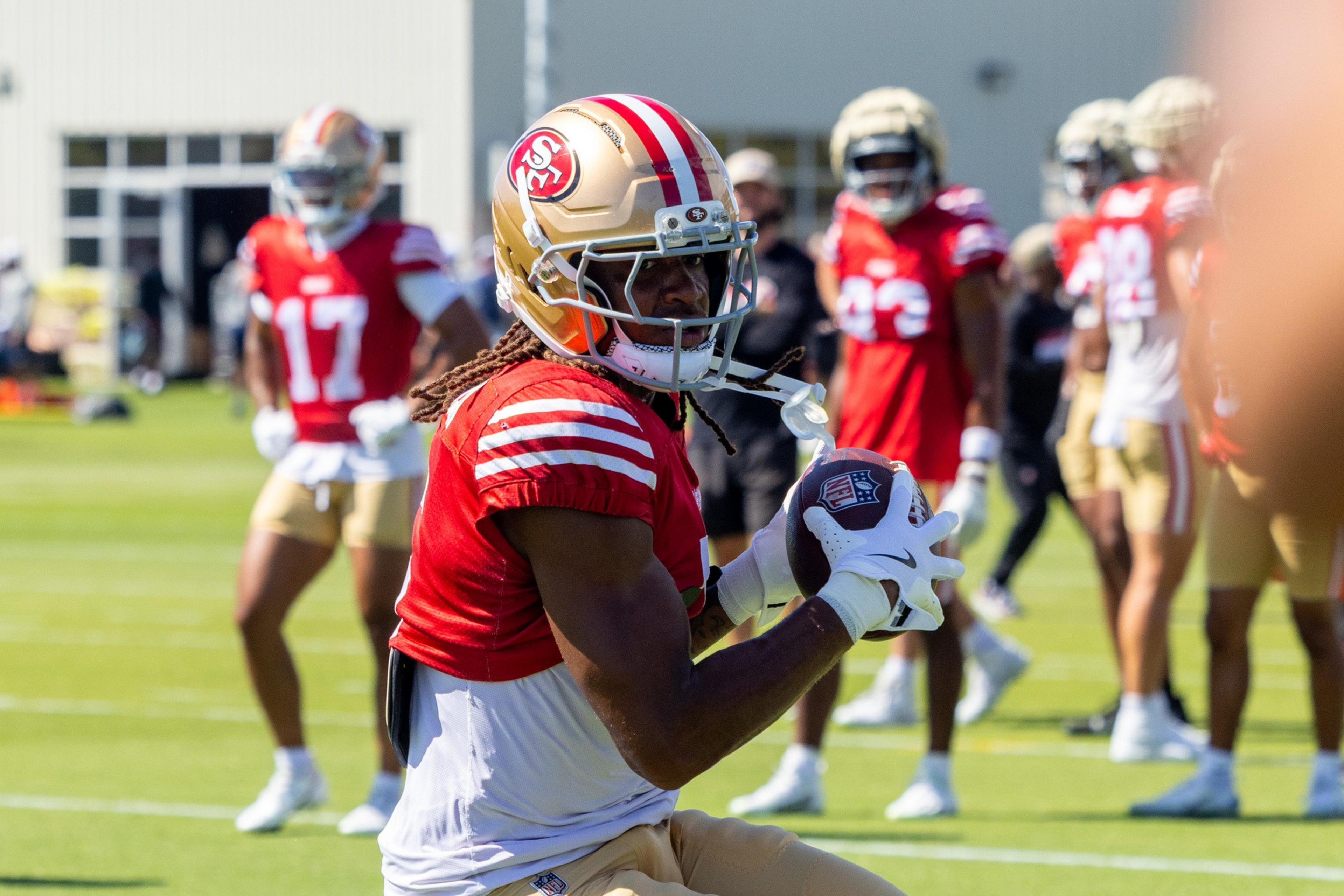 A football player in a red and gold uniform with a helmet holds a football during practice, with teammates in similar uniforms in the background.