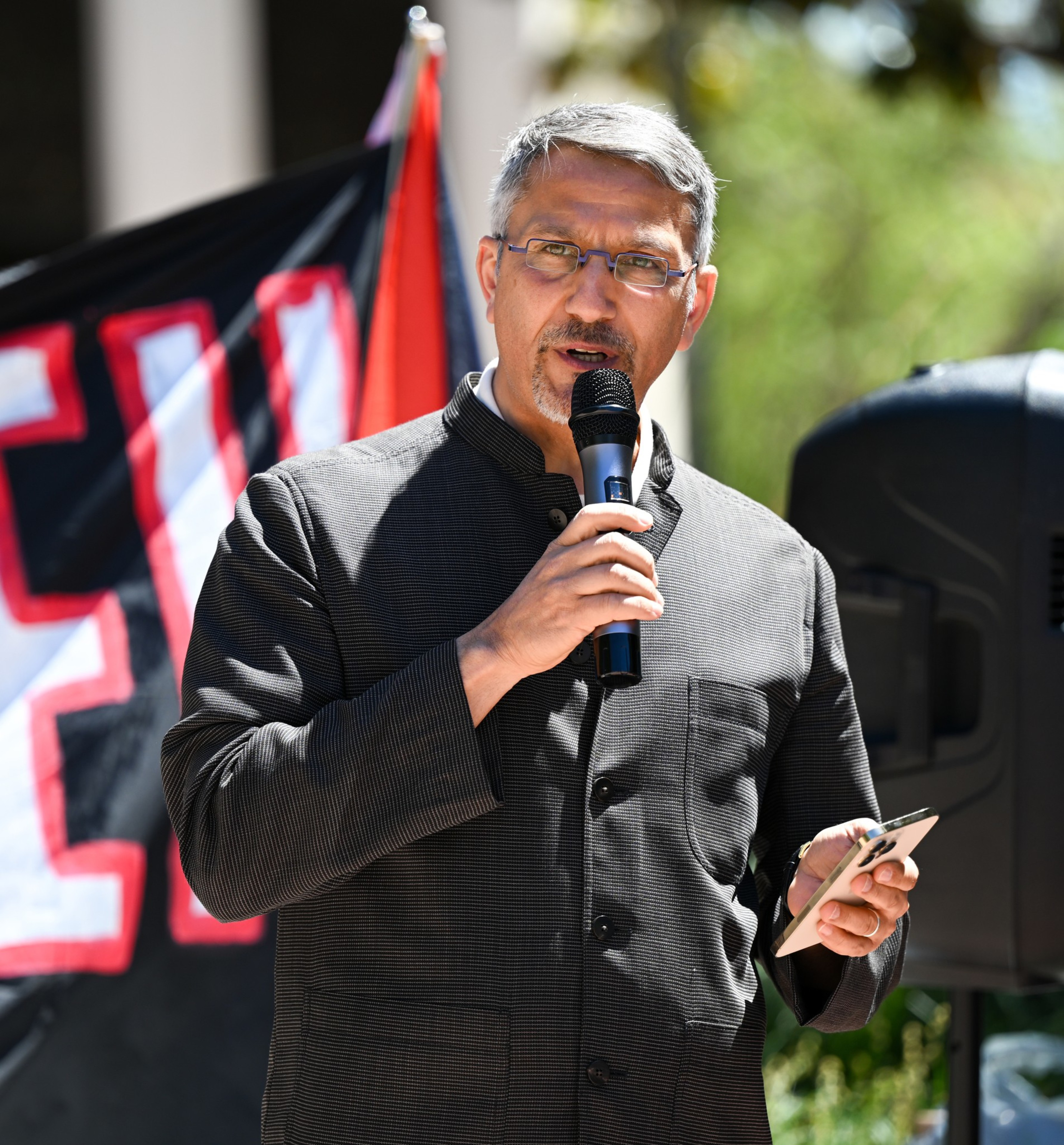 A man is speaking into a microphone at an outdoor event, holding a smartphone, with banners partially visible behind him.