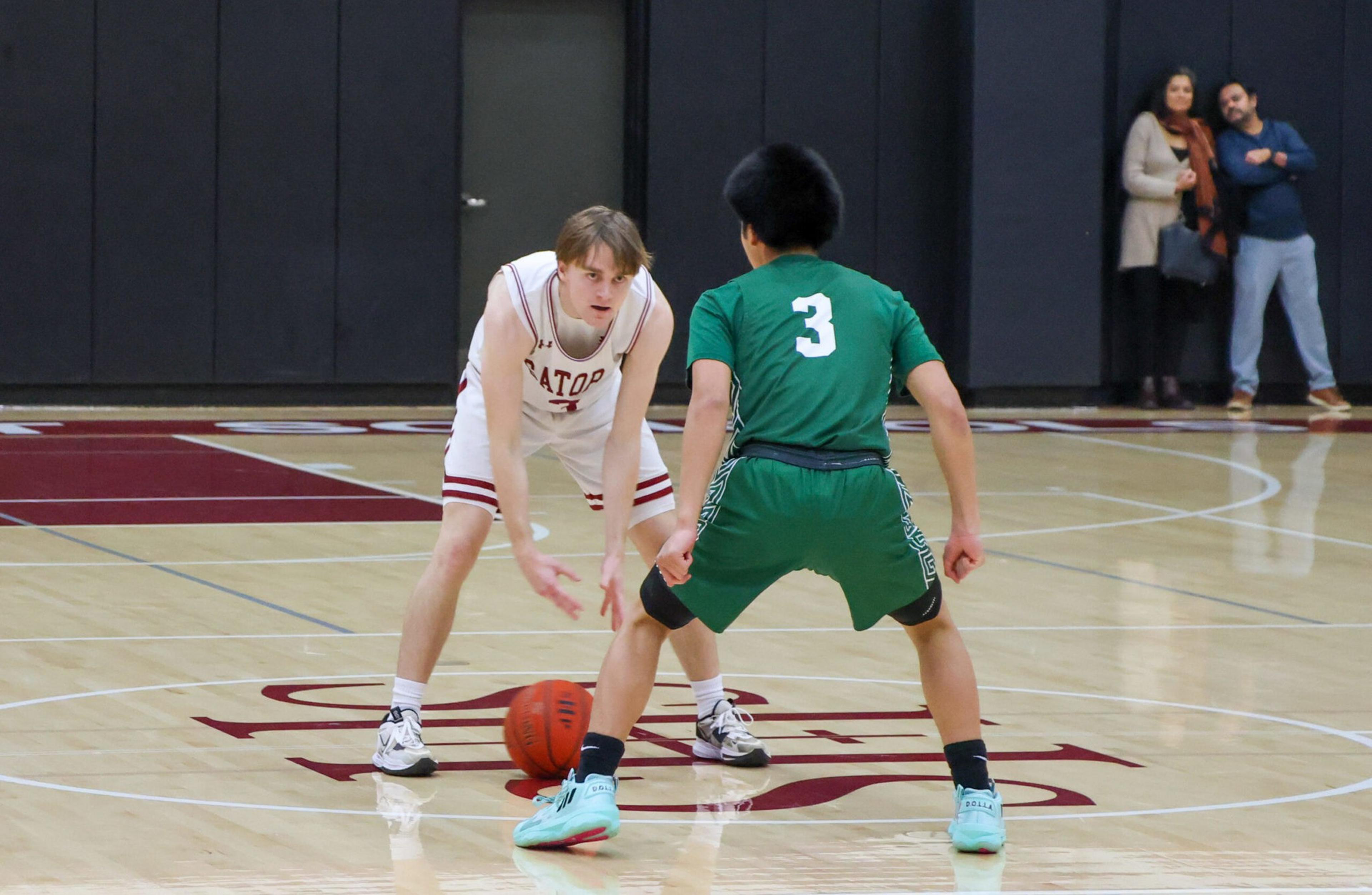 A basketball player in a white uniform dribbles the ball while being guarded by a player in a green uniform with number 3 on his back.