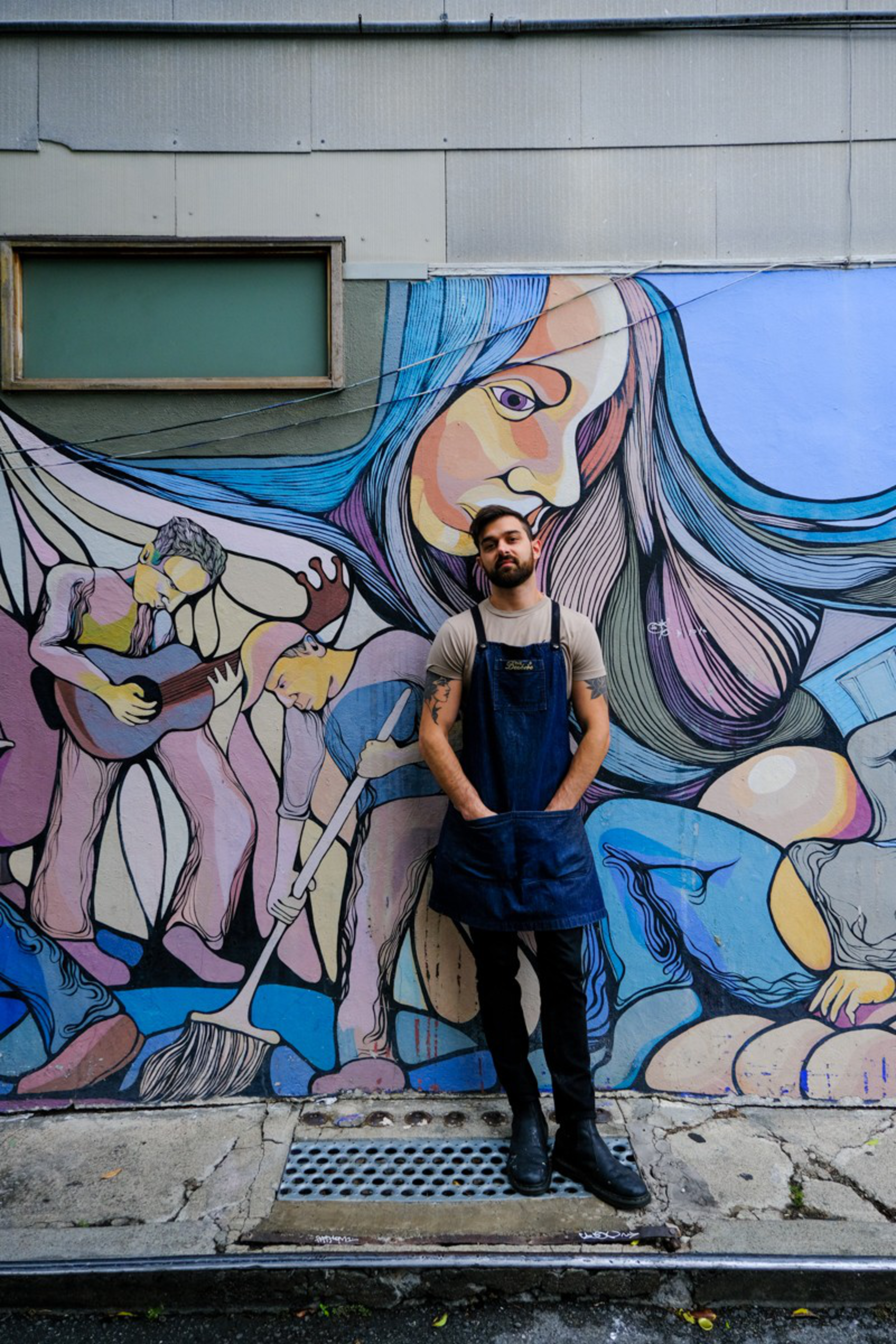 A man with tattoos, wearing a gray shirt and blue apron, stands against a colorful mural depicting musicians and a large face with flowing hair.
