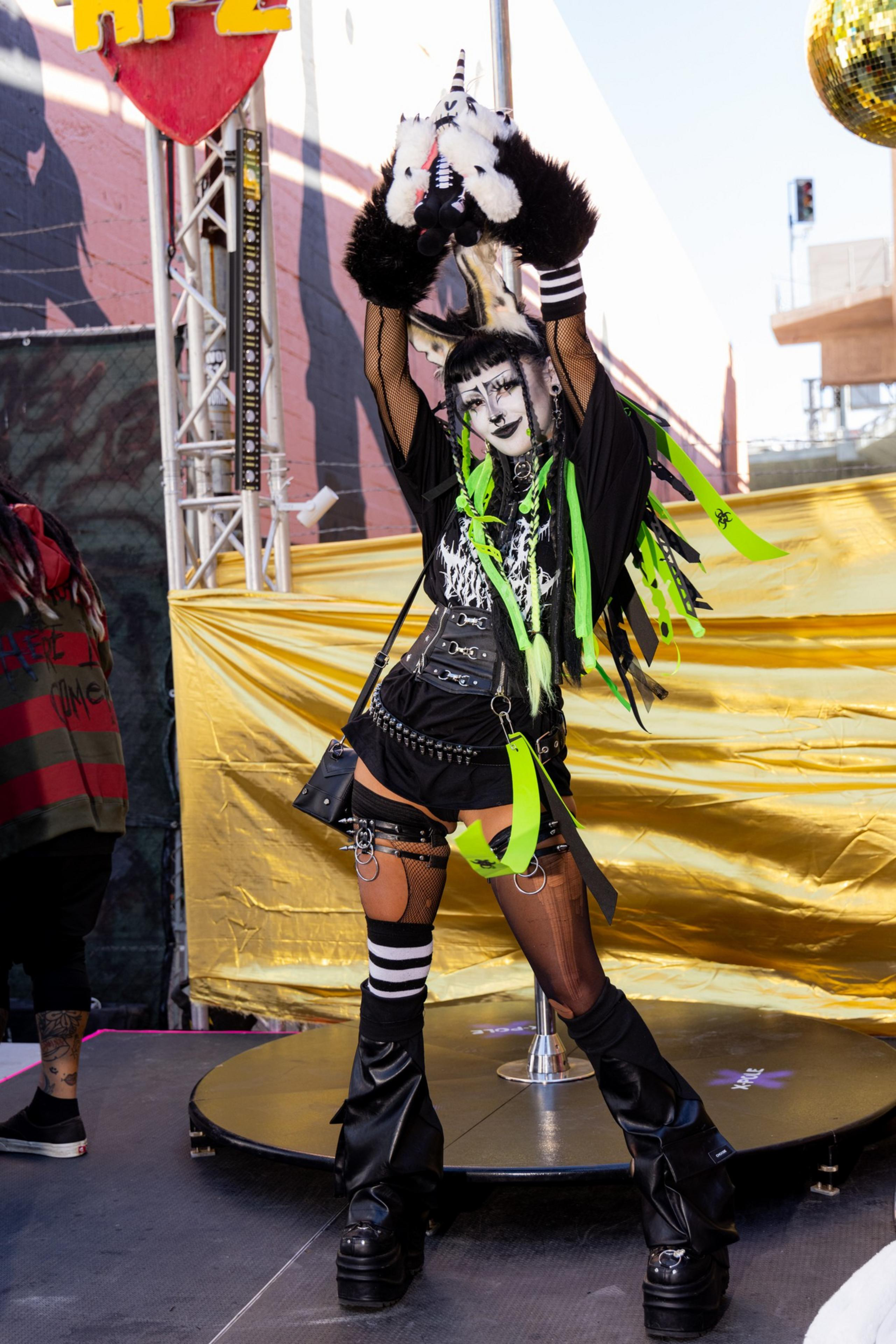 A person in eclectic attire raises a plush toy unicorn aloft. The outfit includes neon green and black elements, chunky platform boots, and bold face makeup.