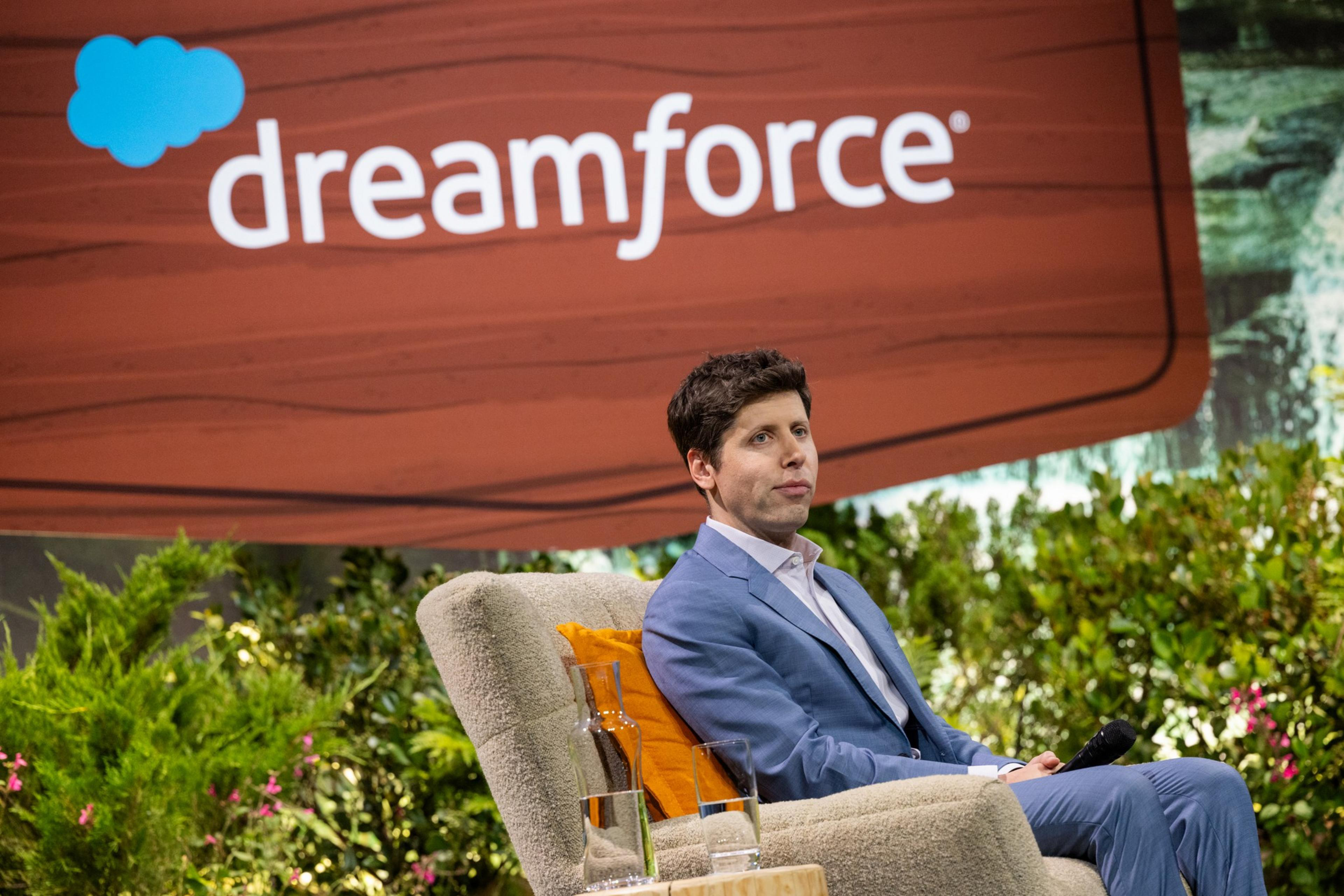A man in a blue suit sits on a beige chair holding a microphone, with a "dreamforce" sign and greenery in the background.
