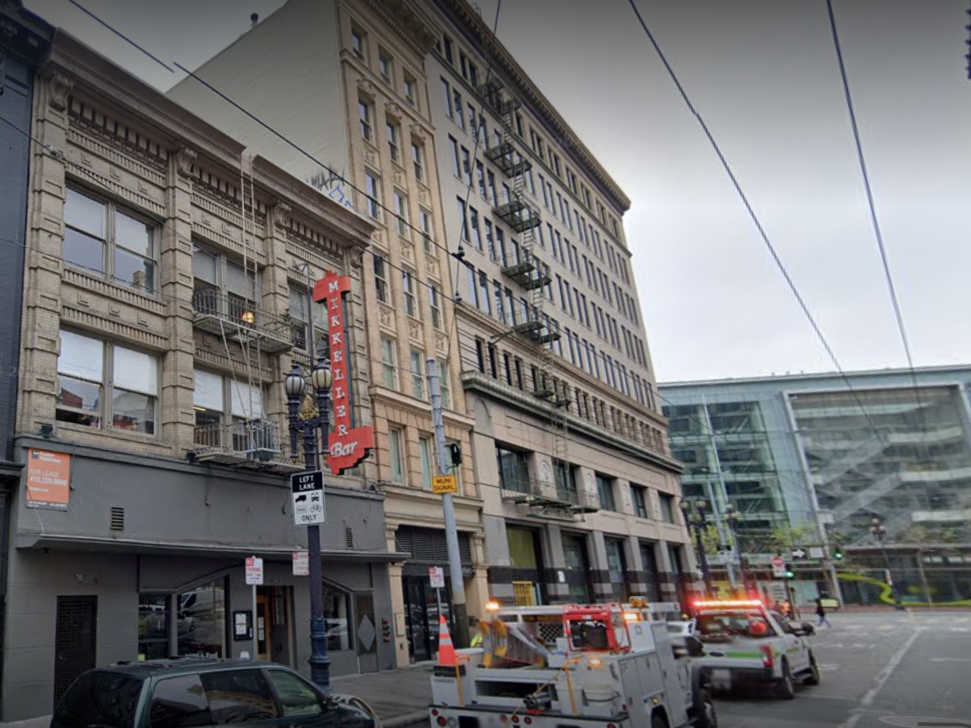 Tall historic buildings with fire escapes line the street, a bright red "Mikkeller Bar" sign hangs, and utility trucks with flashing lights park along the road.