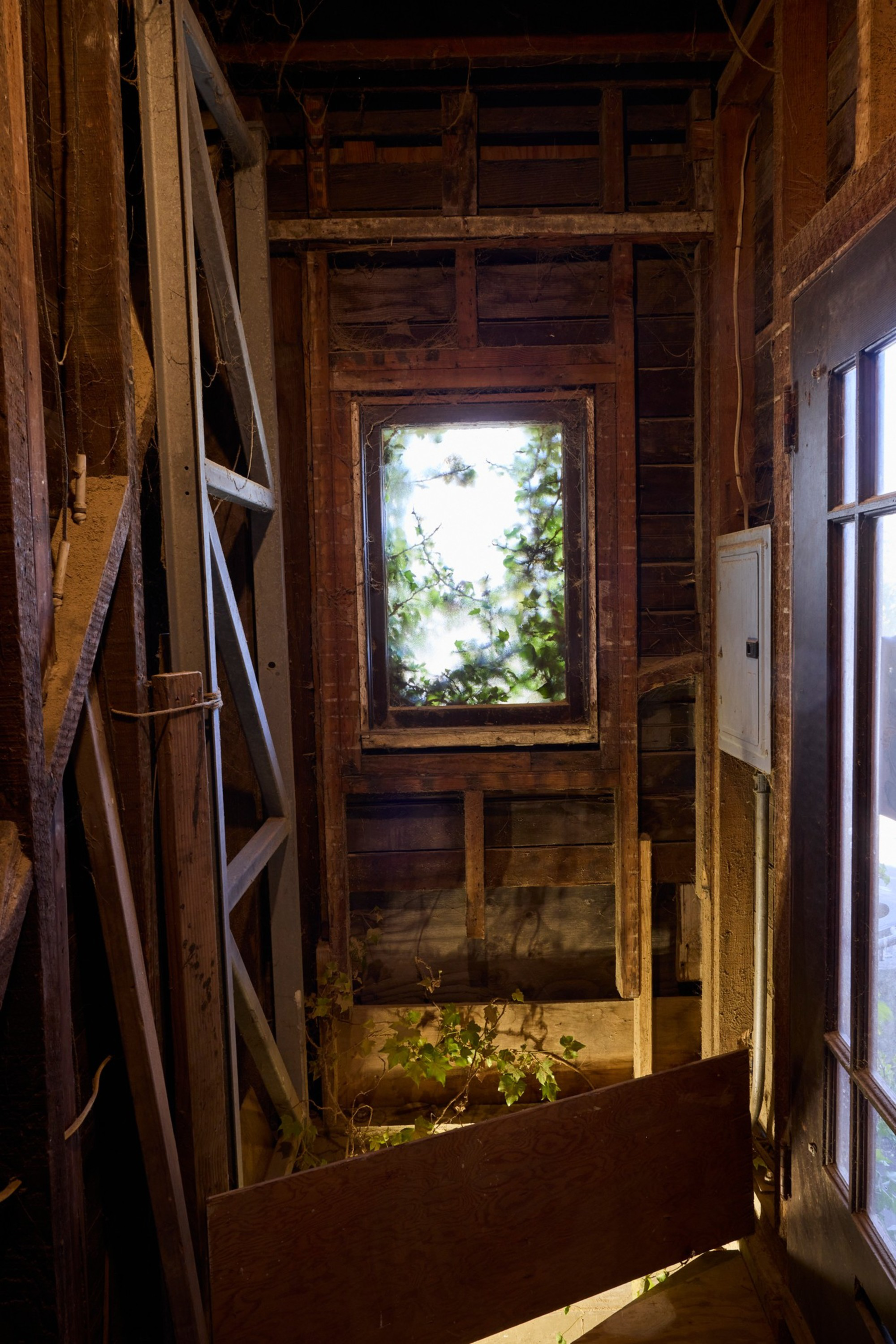An old, unfinished wooden interior space with exposed beams, a small window showing greenery outside, a leaning ladder, and a partially blocking wooden board.