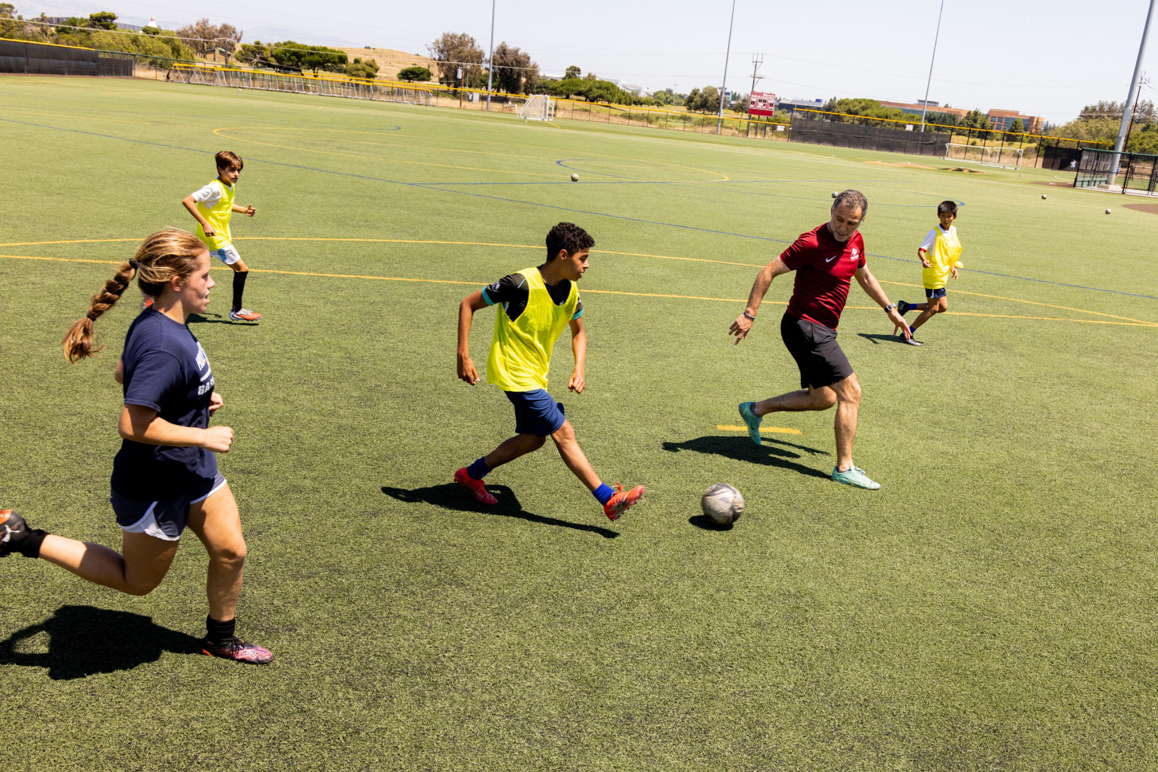 A group of kids and an adult man play soccer on a sunny field, with some wearing yellow practice vests and actively chasing the ball.