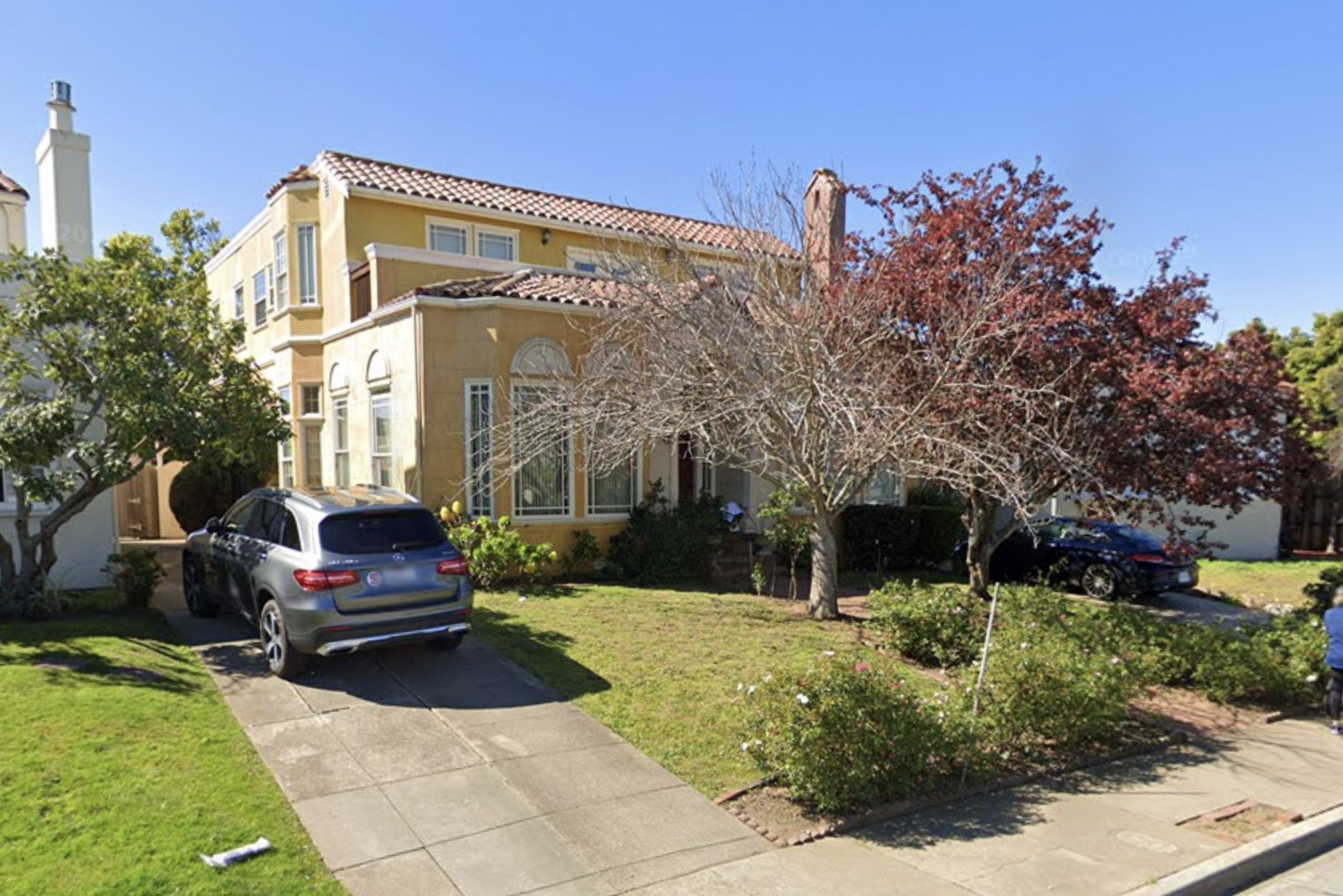 A two-story yellow house with a tile roof, driveway, green lawn, and a silver SUV parked outside.