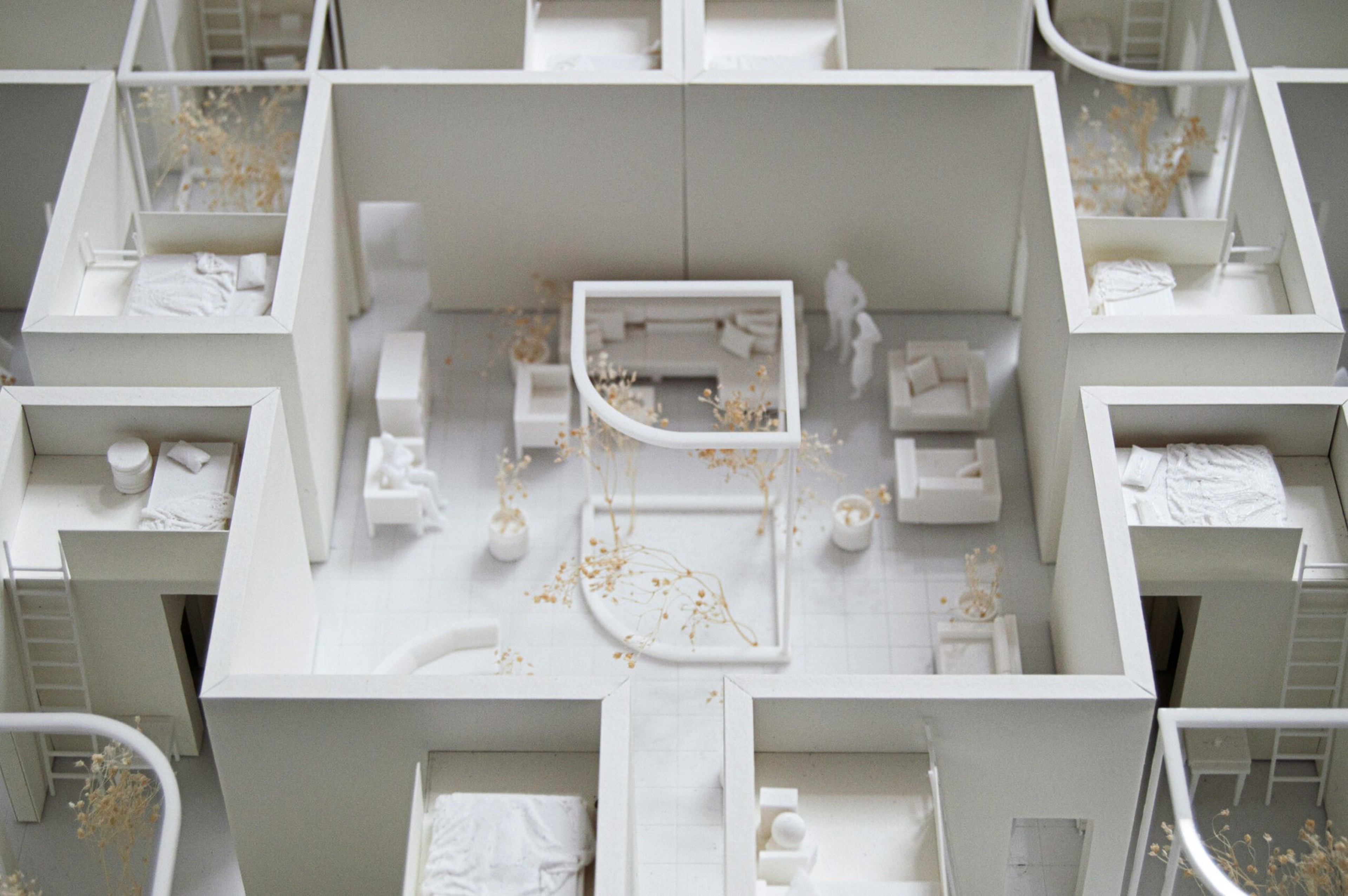 A white architectural model depicts a floor plan with multiple rooms, beds, couches, small plants, and two human figures in a central living area.