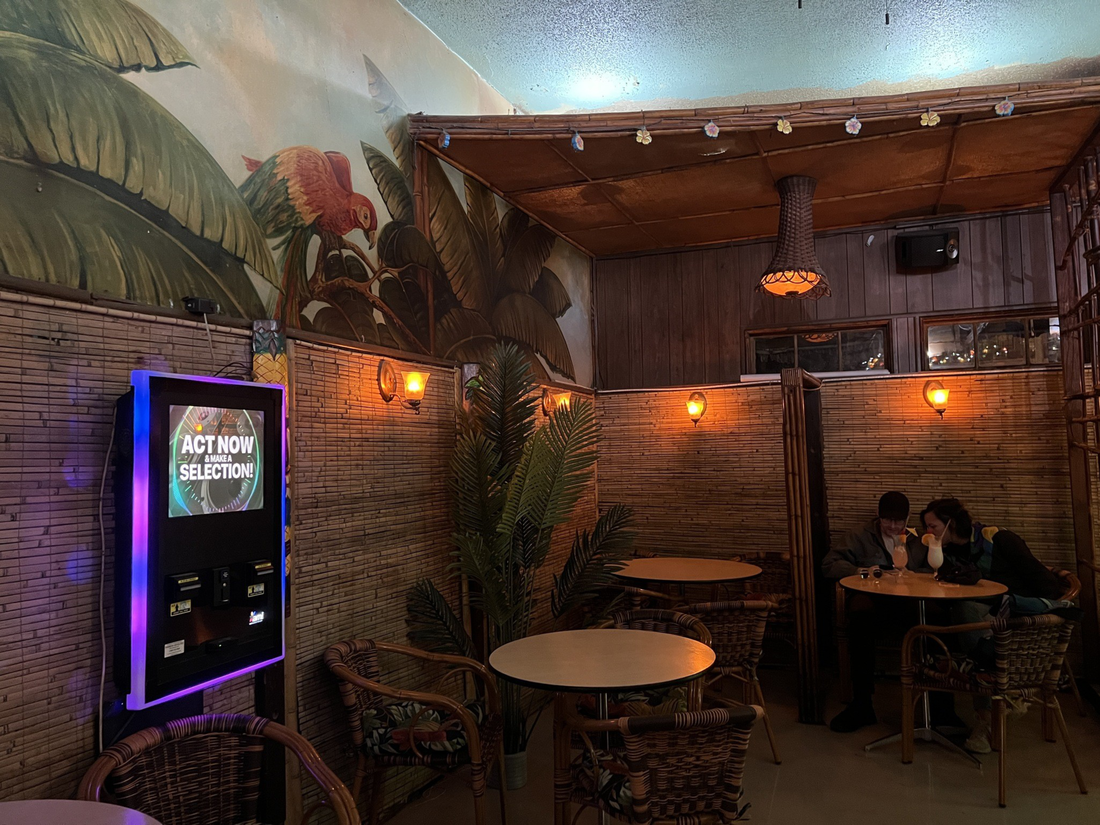 Two customers sit at a round table inside a bar that has two other round tables show and a jukebox machine hanging from the wall. 