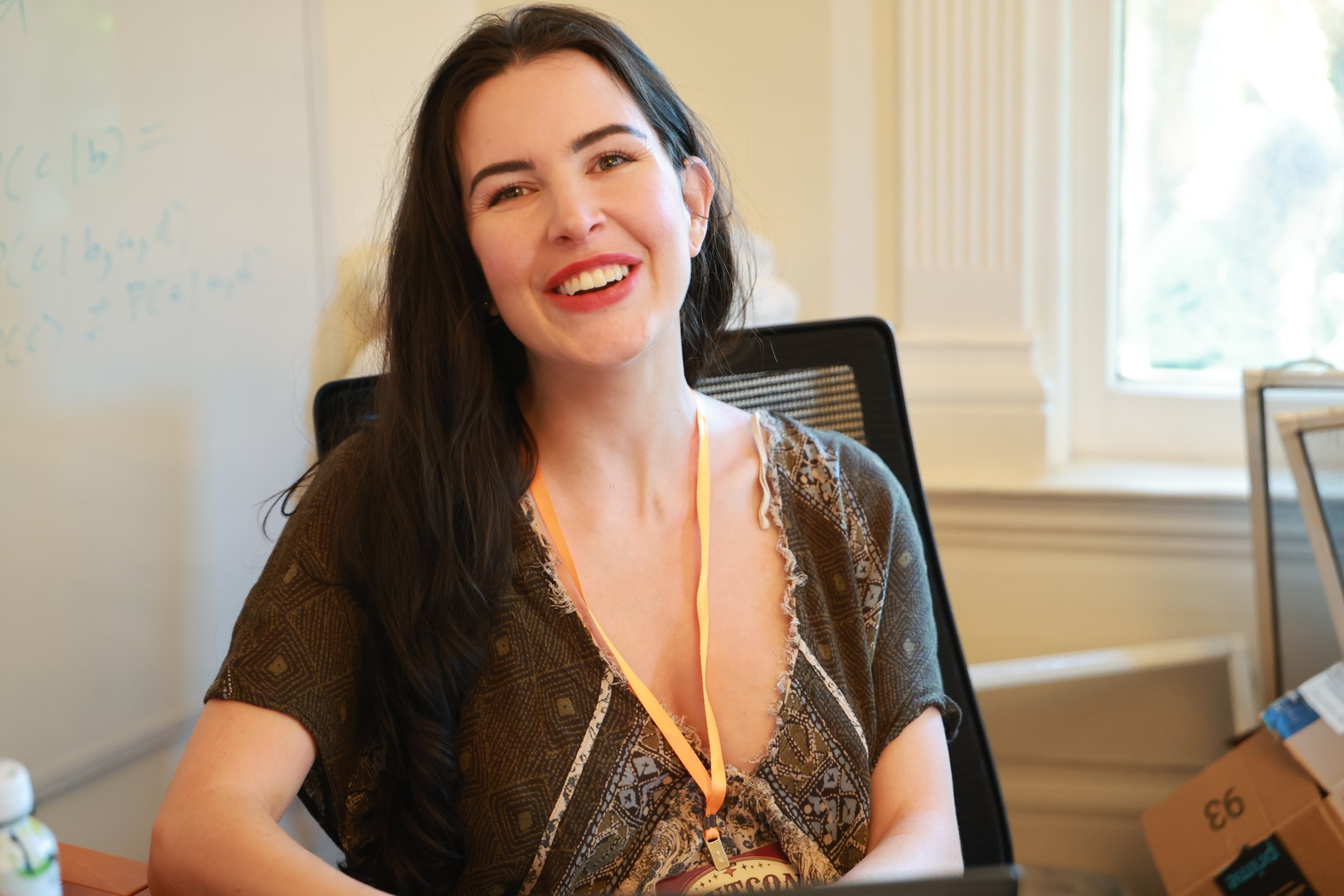 A woman with long dark hair wearing a patterned top and an orange lanyard smiles while sitting on an office chair near a window.