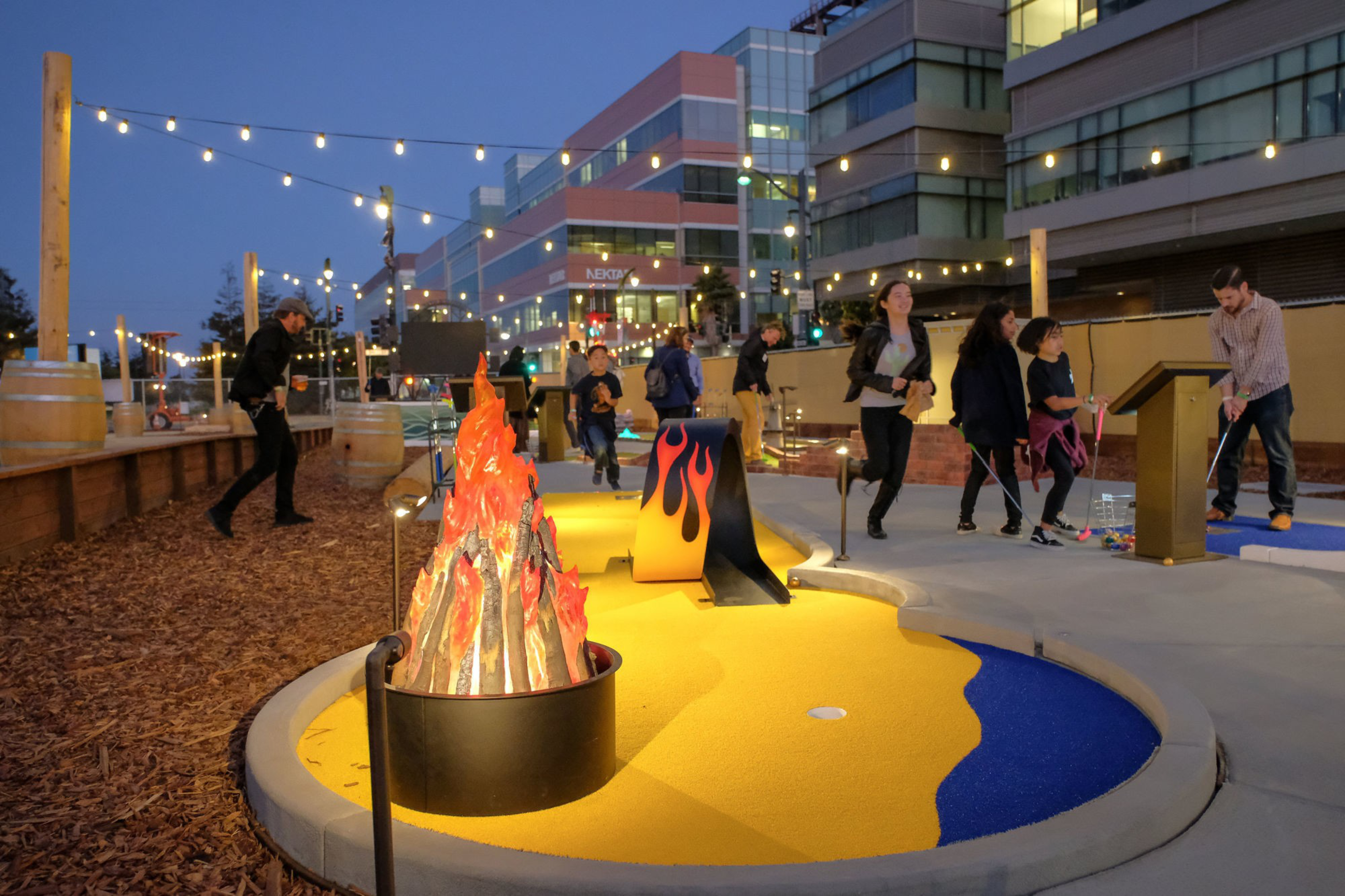 People play miniature golf around a fake plastic fire pit under strings of lights.