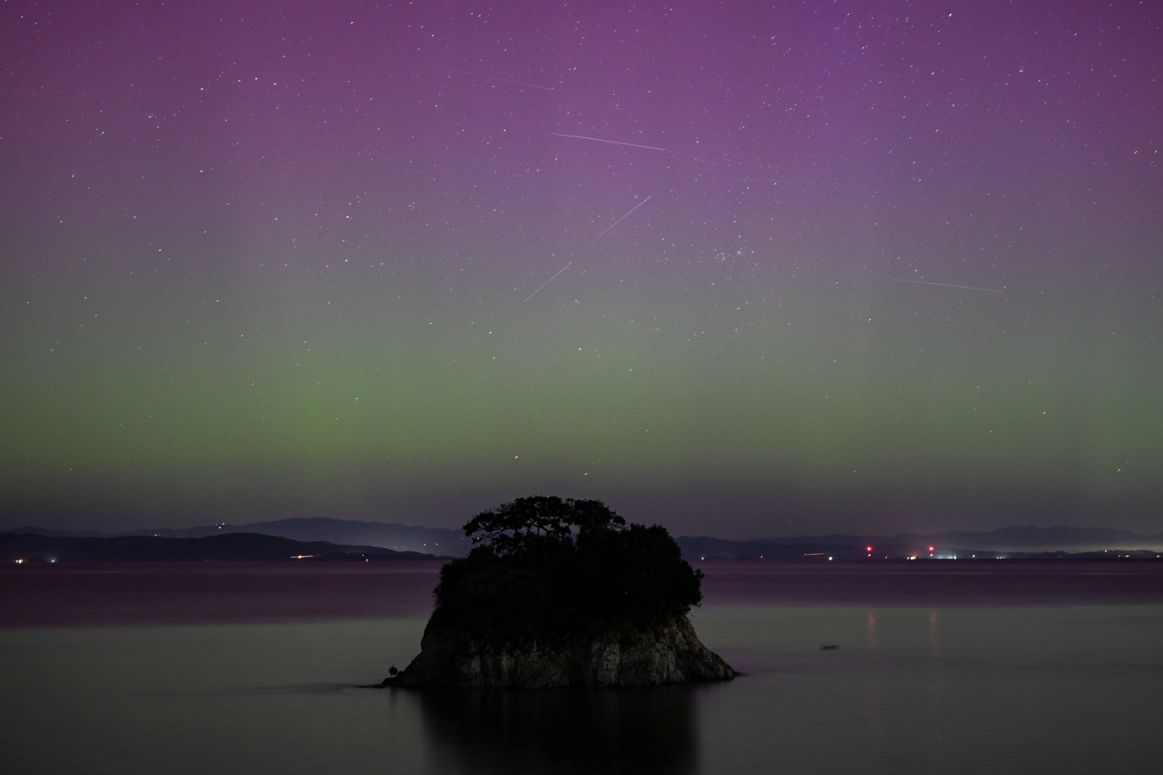 A small island with trees sits in calm water under a night sky with stars and aurora-like lights.