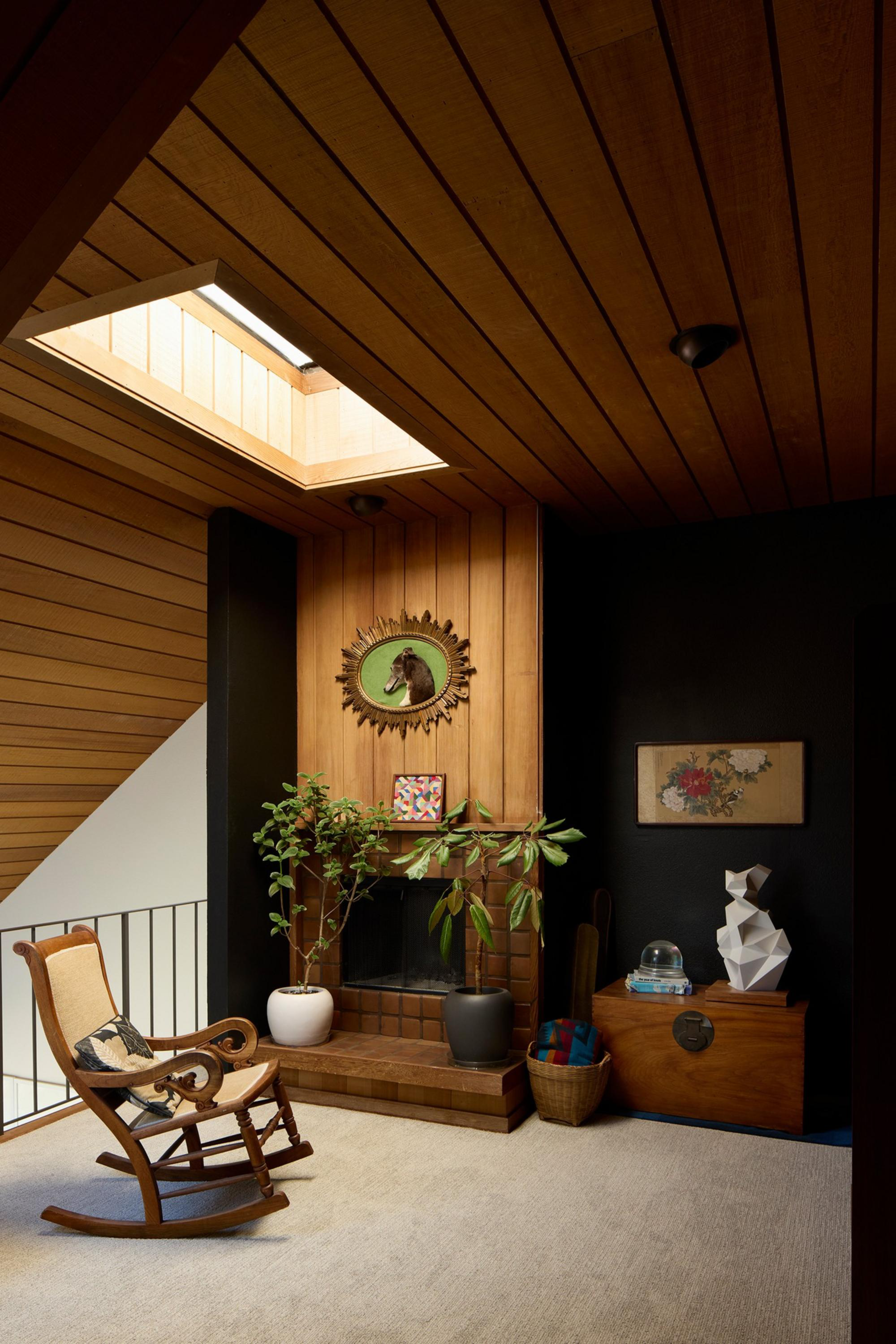 A cozy corner with wood-paneled walls and ceiling, featuring a rocking chair, potted plants, a fireplace, and art on the walls.