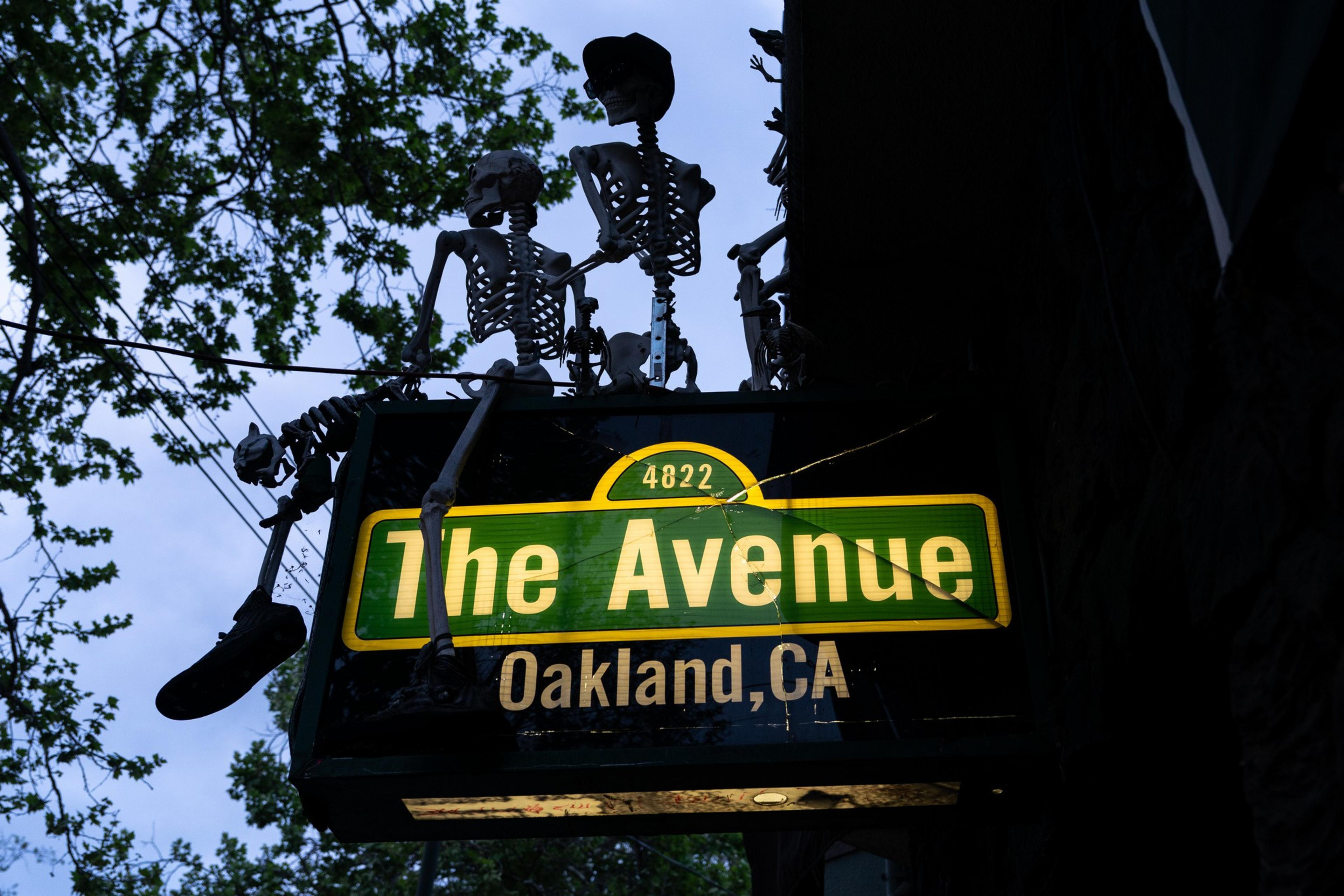 A neon sign for &quot;The Avenue&quot; in Oakland, CA, is topped by skeleton decorations. The sign is cracked, and the surroundings have tree branches.