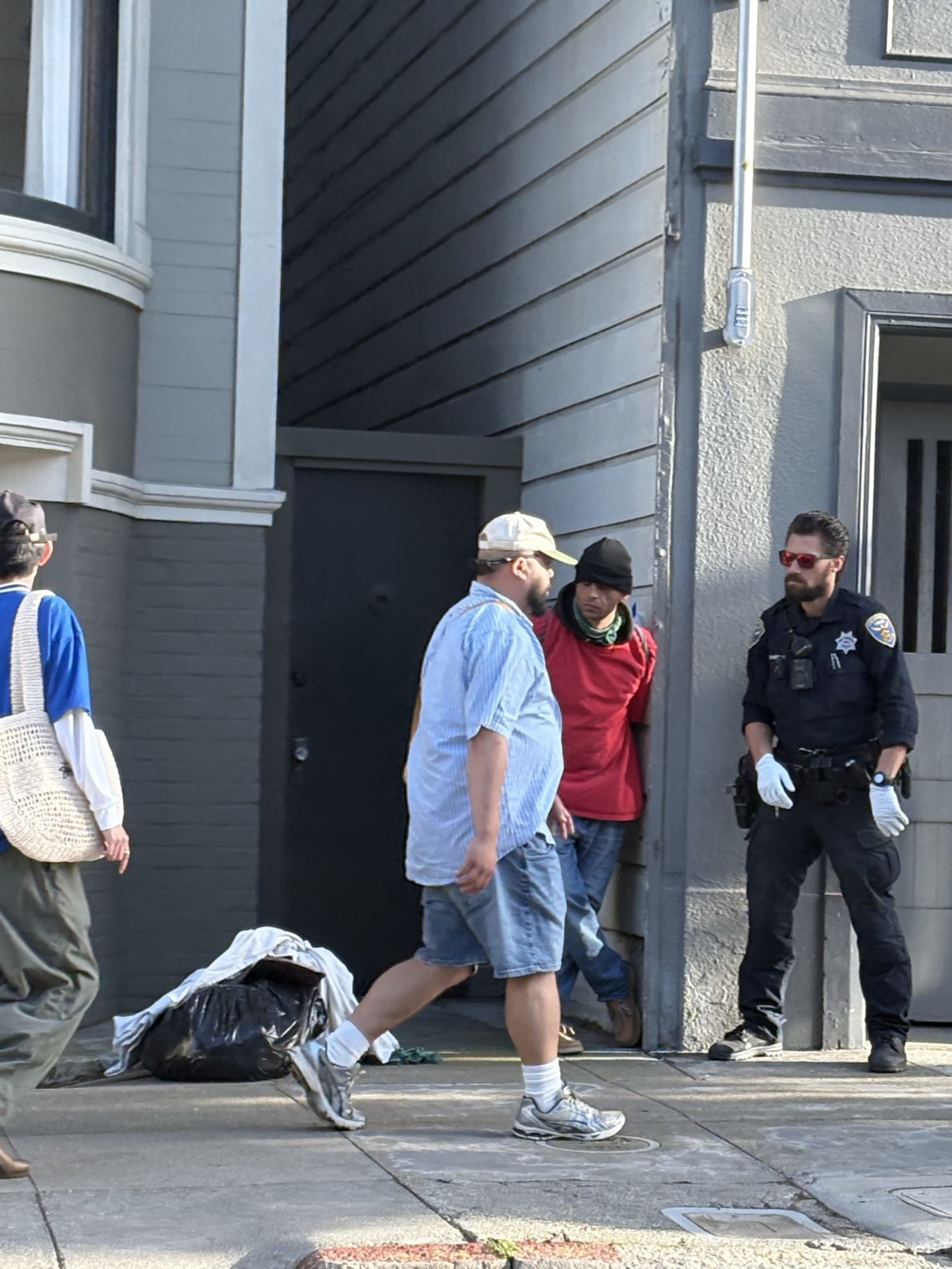 A police officer with gloves stands near a person in a red hoodie against a wall, while others walk by on the sidewalk, with bags and items nearby.