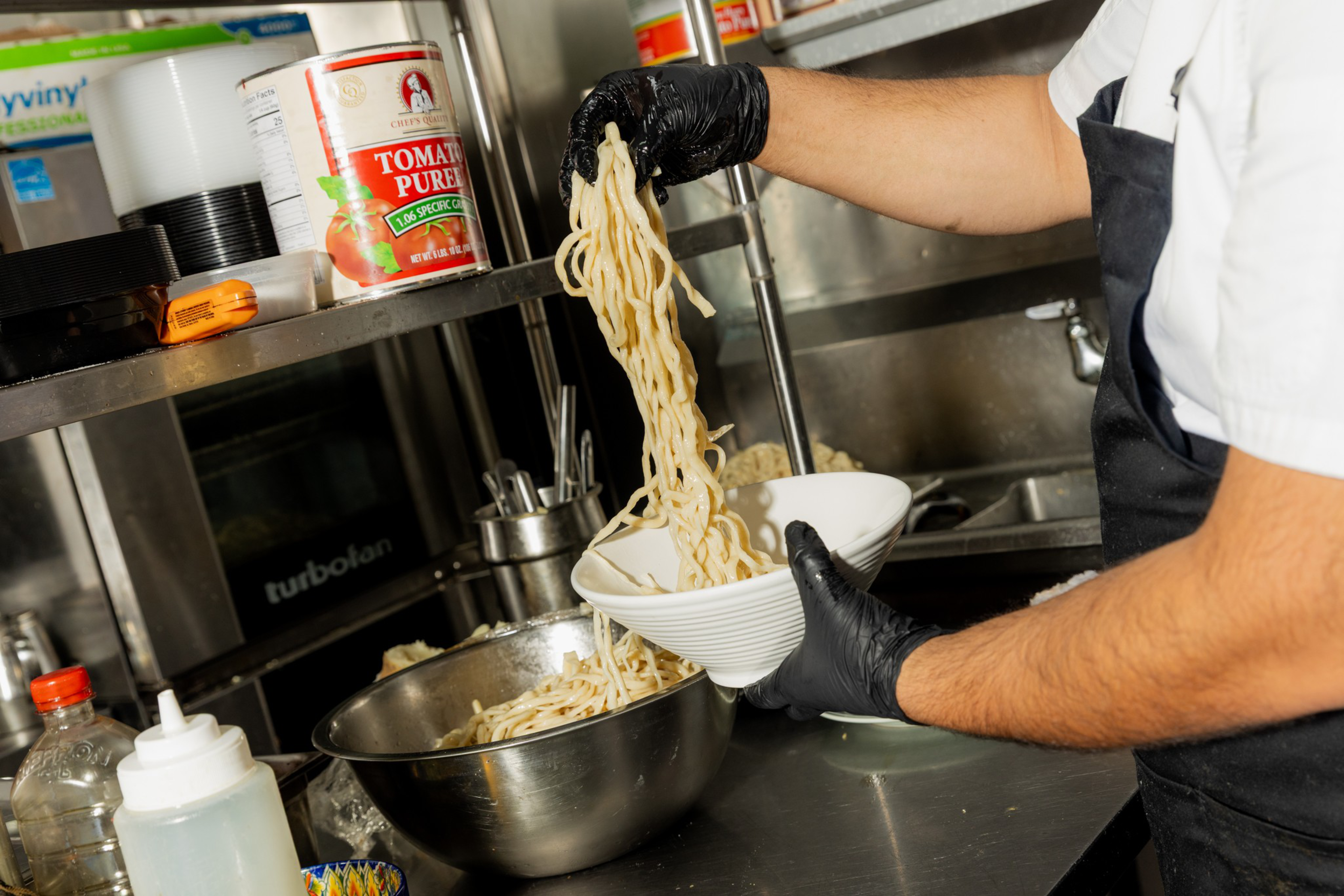A person wearing black gloves transfers noodles from a large metal bowl into a white bowl in a kitchen setting.