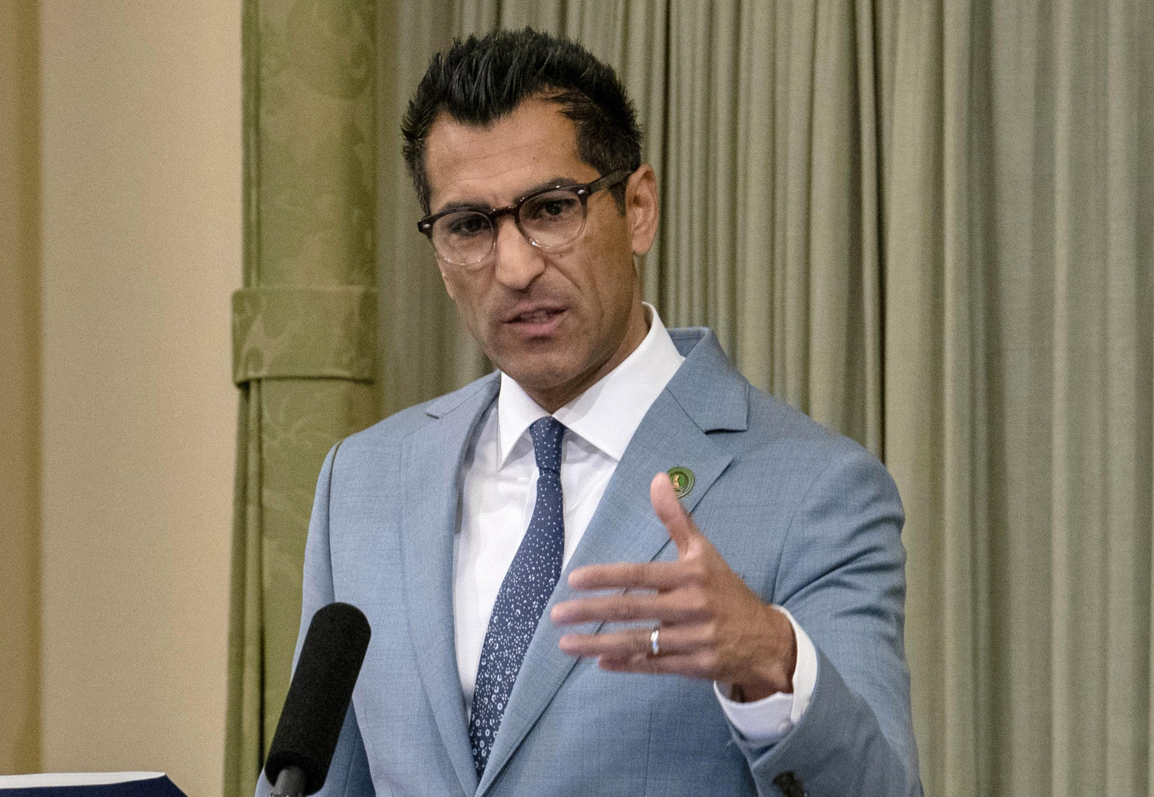 A man in a light blue suit and glasses gestures while speaking at a podium.