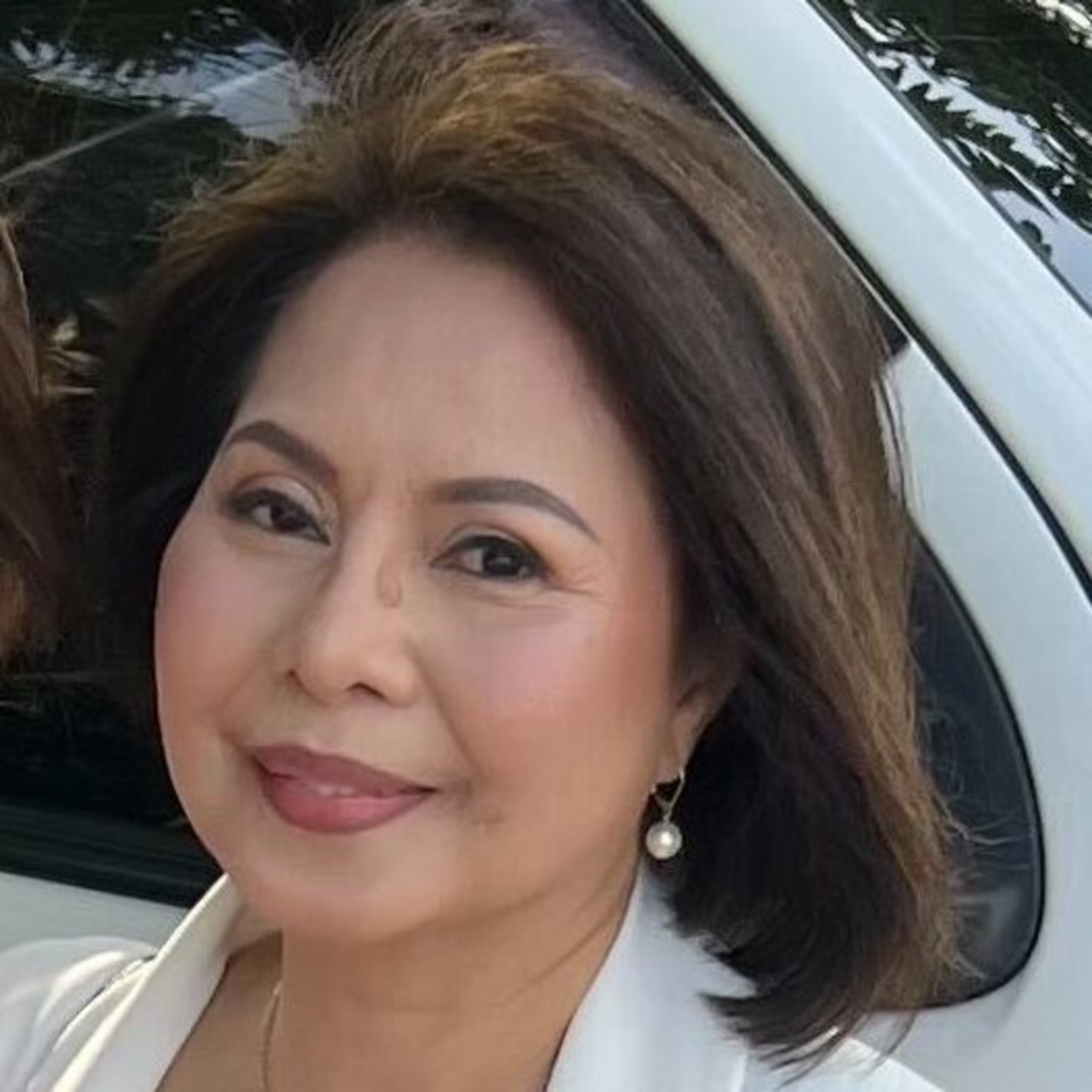 A woman with short, dark hair and makeup smiles while wearing a pearl earring and a white top. She stands in front of a white vehicle with trees reflecting on its windows.