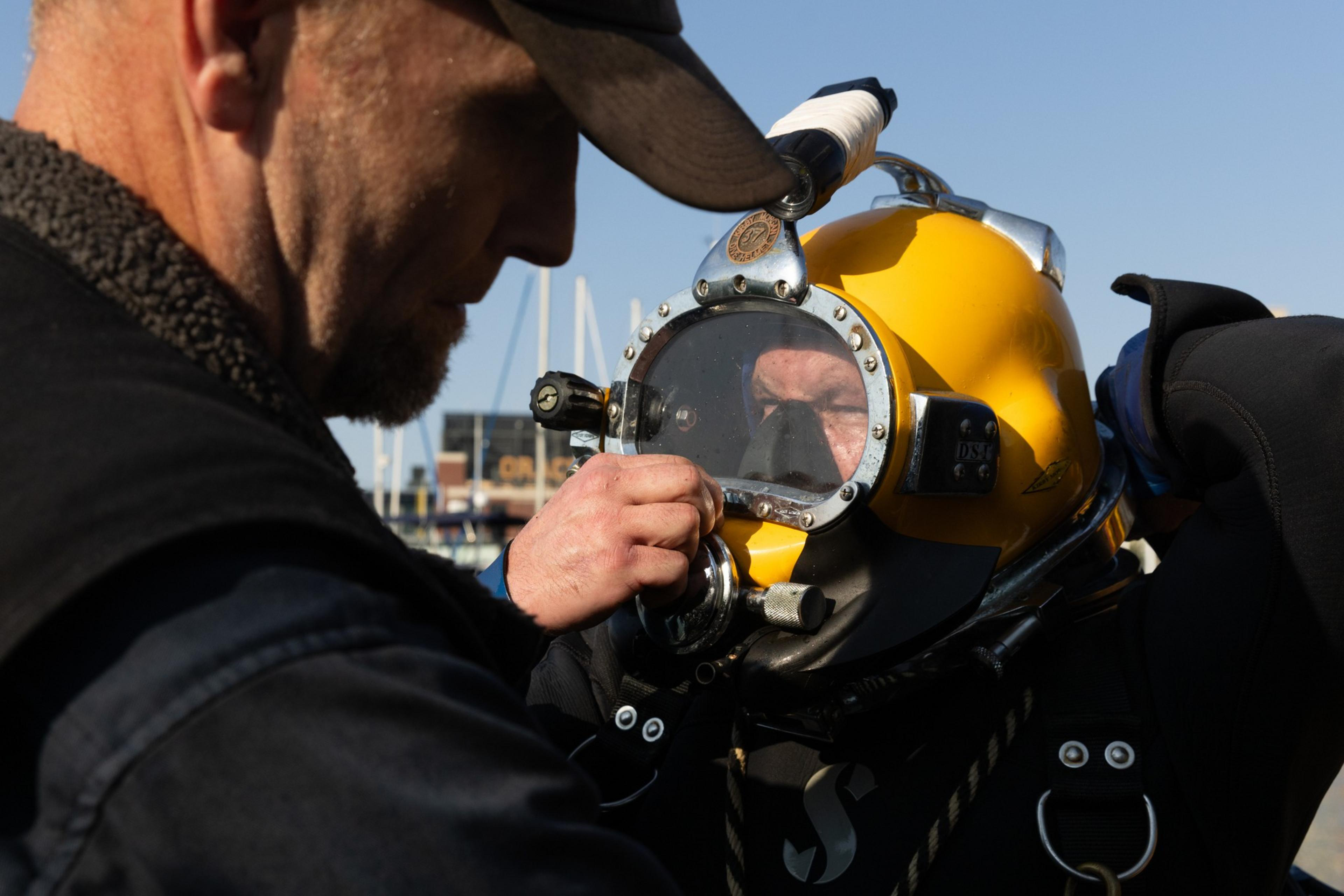 A man helps another person adjust a yellow diving helmet with a metal frame and glass faceplate, both dressed in wetsuits near a harbor.