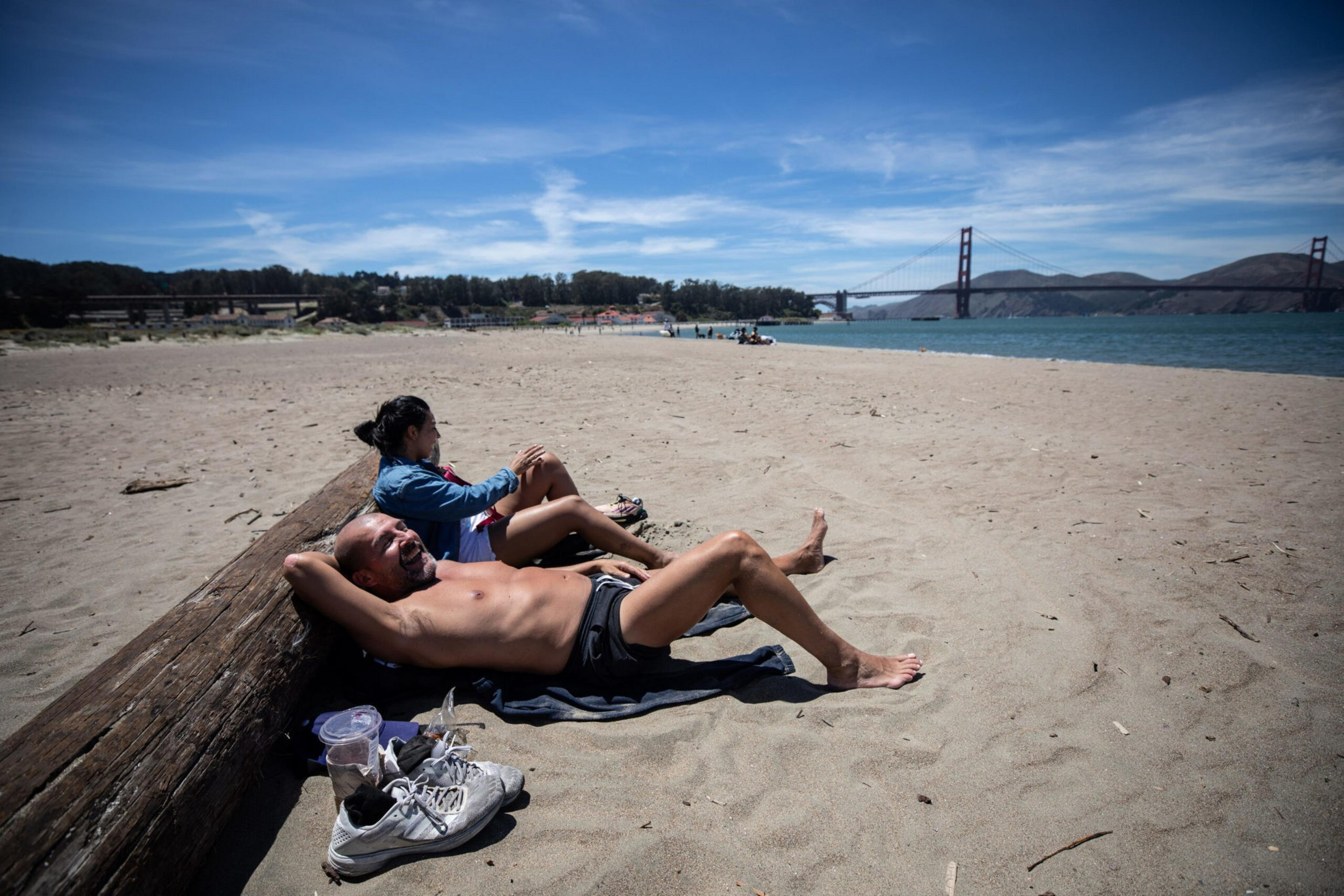 A shirtless man and a woman sit and lie on a sandy beach near a large log, with the Golden Gate Bridge visible in the distance.