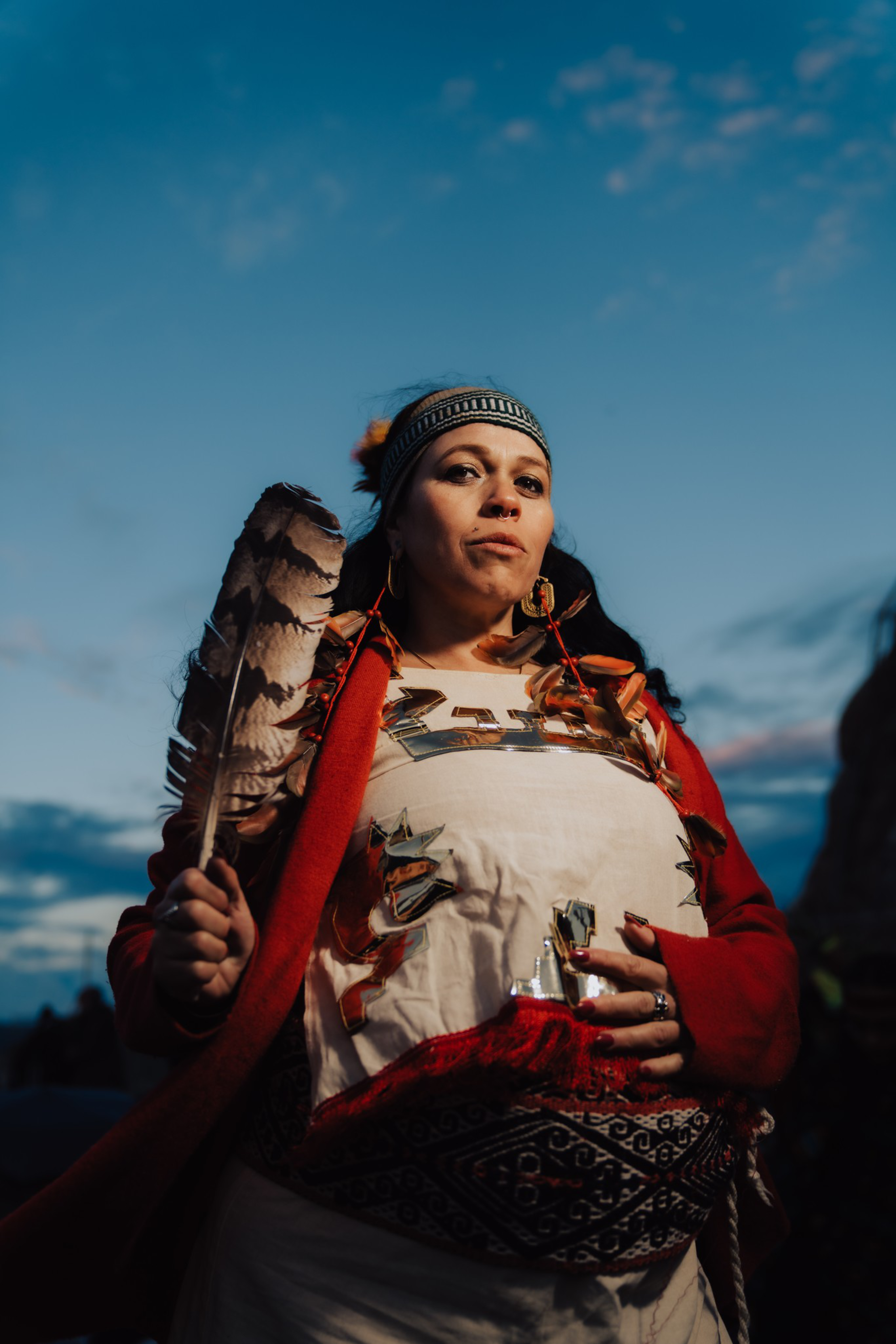 A woman wearing traditional attire holds a large feather, with a patterned headband and a red garment against a dusk sky.