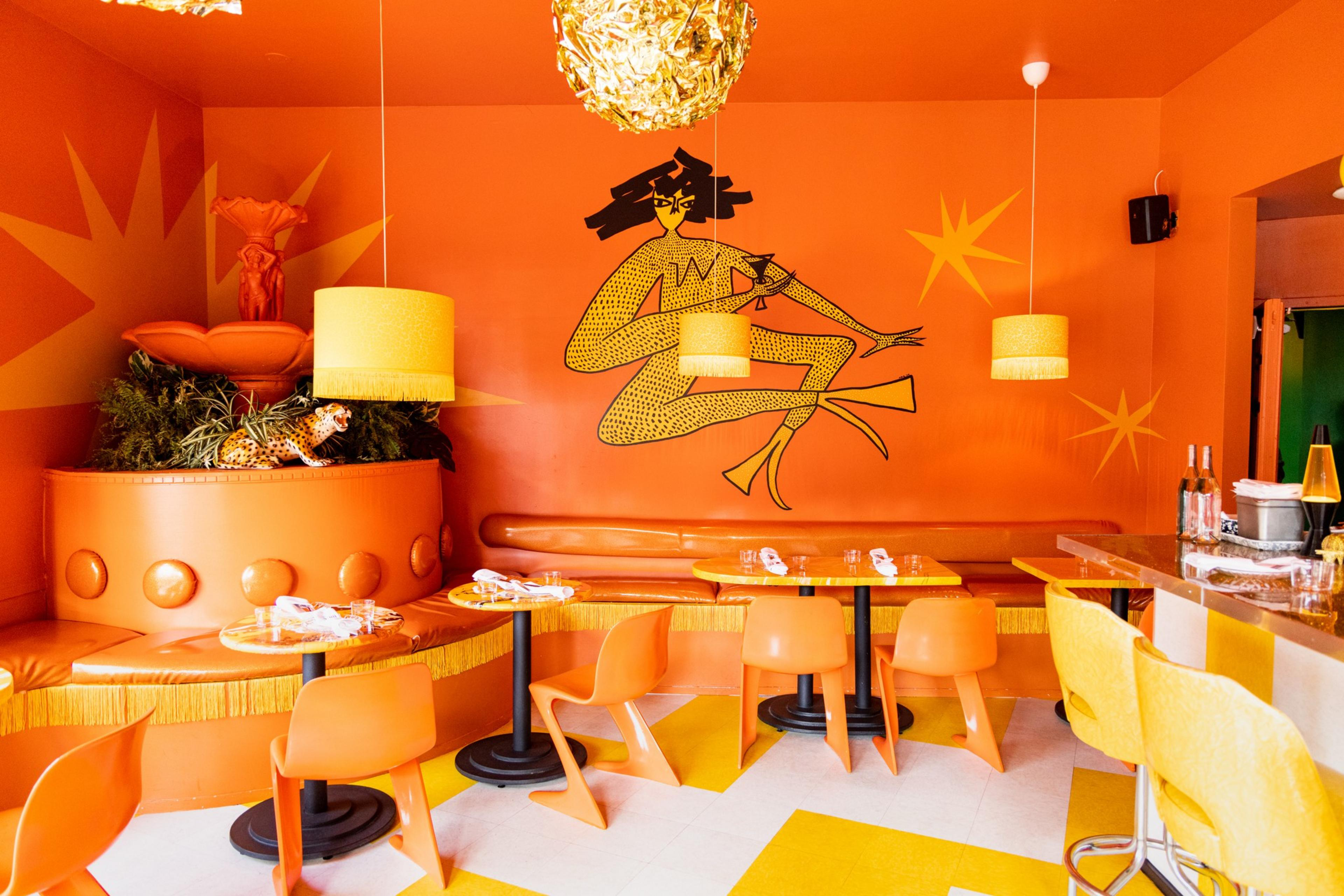 A vibrant orange-themed room with retro orange chairs, a curved bench, yellow pendant lights, a bar with yellow stools, and a large wall mural of a stylized figure.
