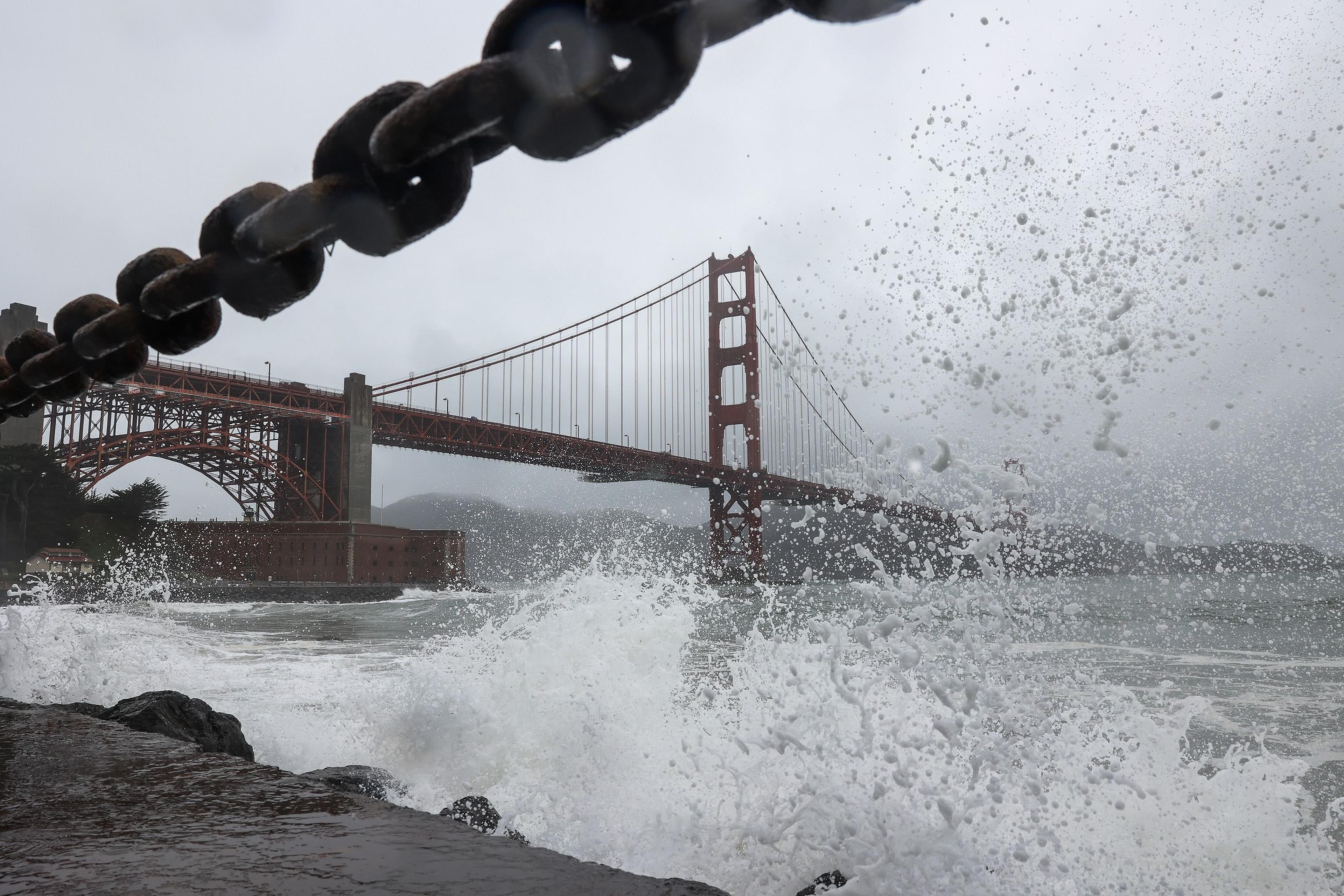 The Golden Gate Bridge looms behind a spray of sea foam, partially obscured by a large, rusty chain in the foreground.