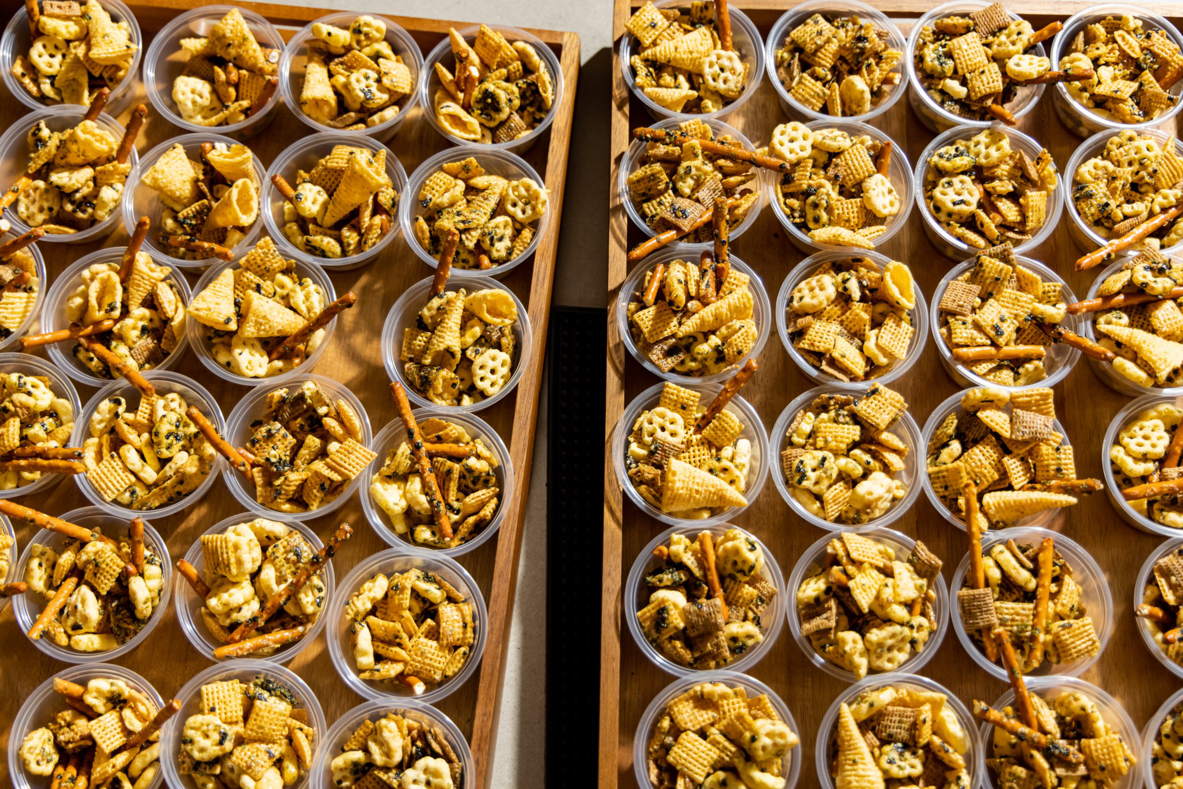 Multiple small clear cups filled with assorted snack mix, including pretzel sticks, crunchy cereals, and cheese crackers, arranged neatly on wooden trays.