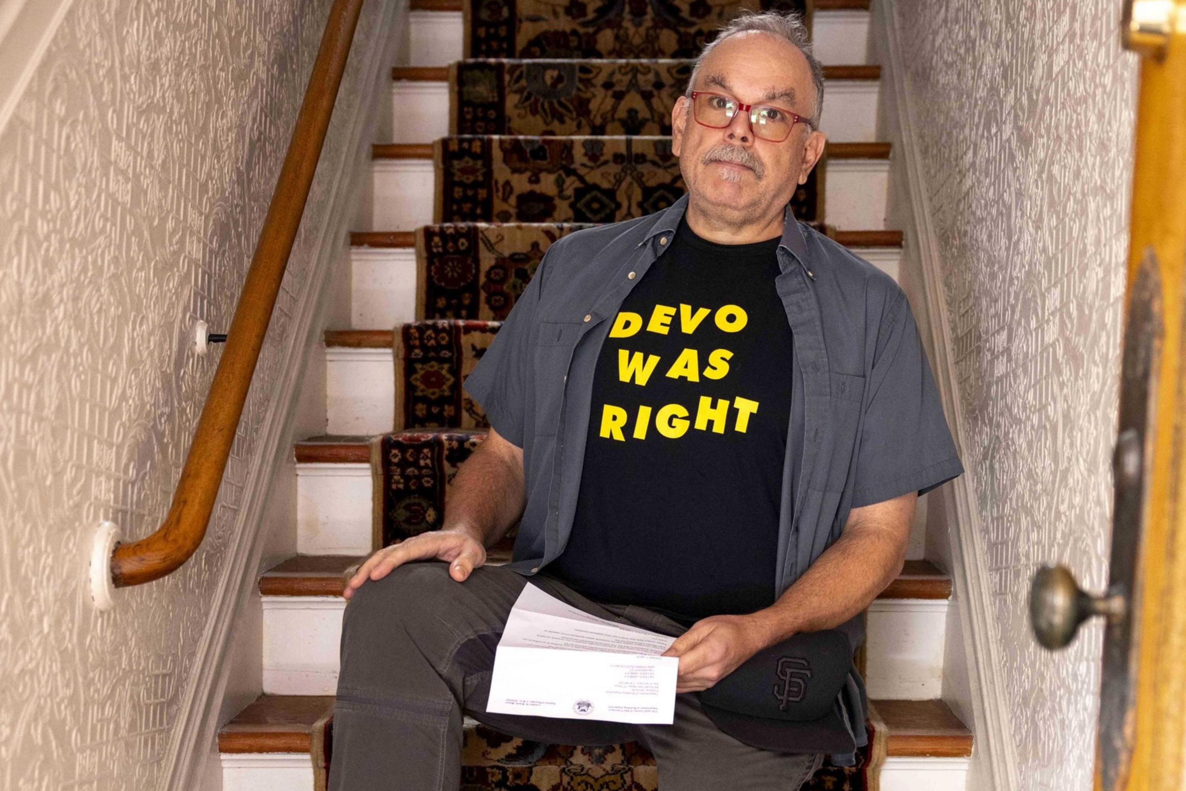 A person sits on a patterned staircase wearing a "DEVO WAS RIGHT" t-shirt, gray pants, and black shoes, holding a piece of paper, with a wooden handrail beside them.
