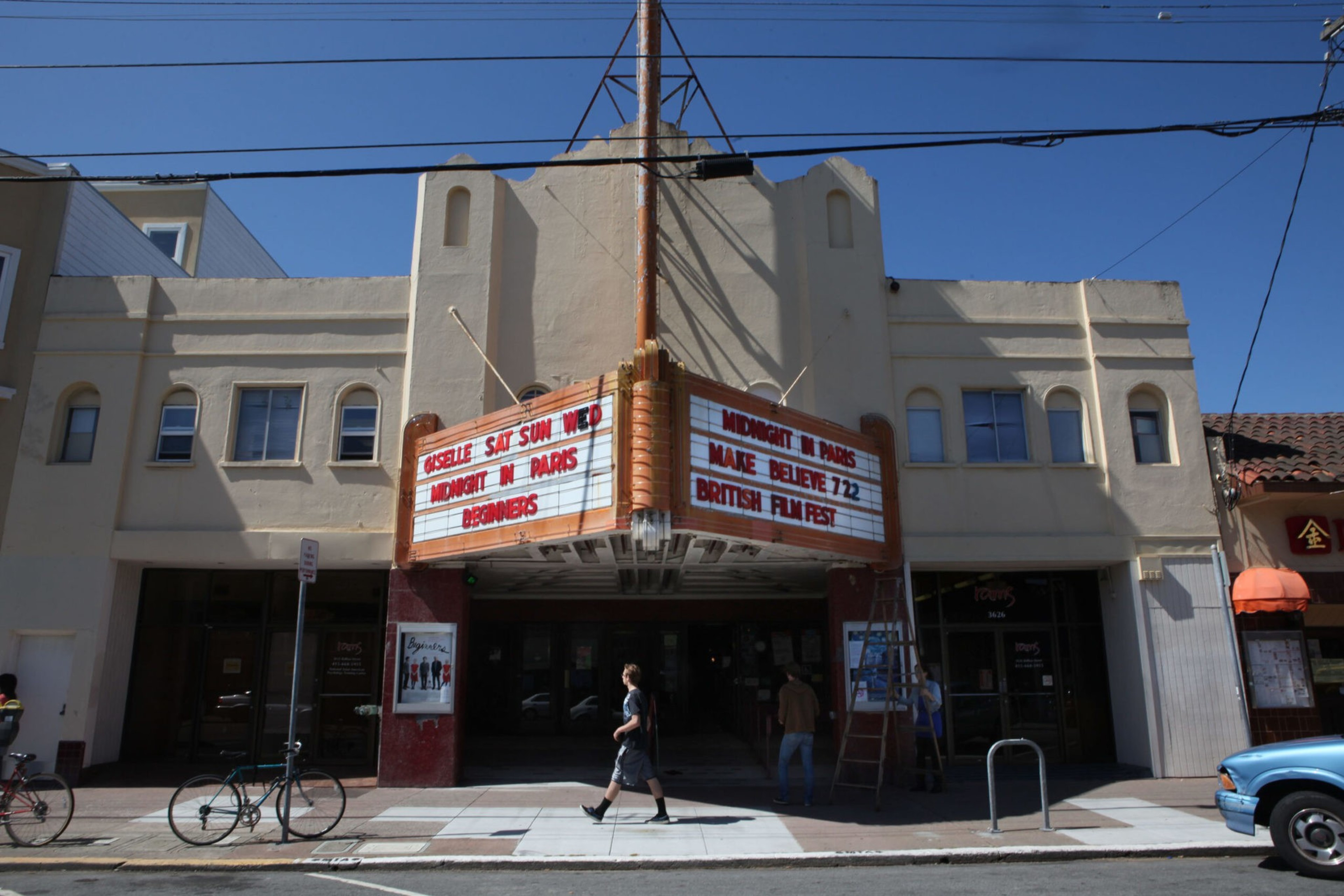 A vintage movie theater with a beige facade displays films "Midnight in Paris" and "Make Believe" on a marquee, while people walk past on the sidewalk.