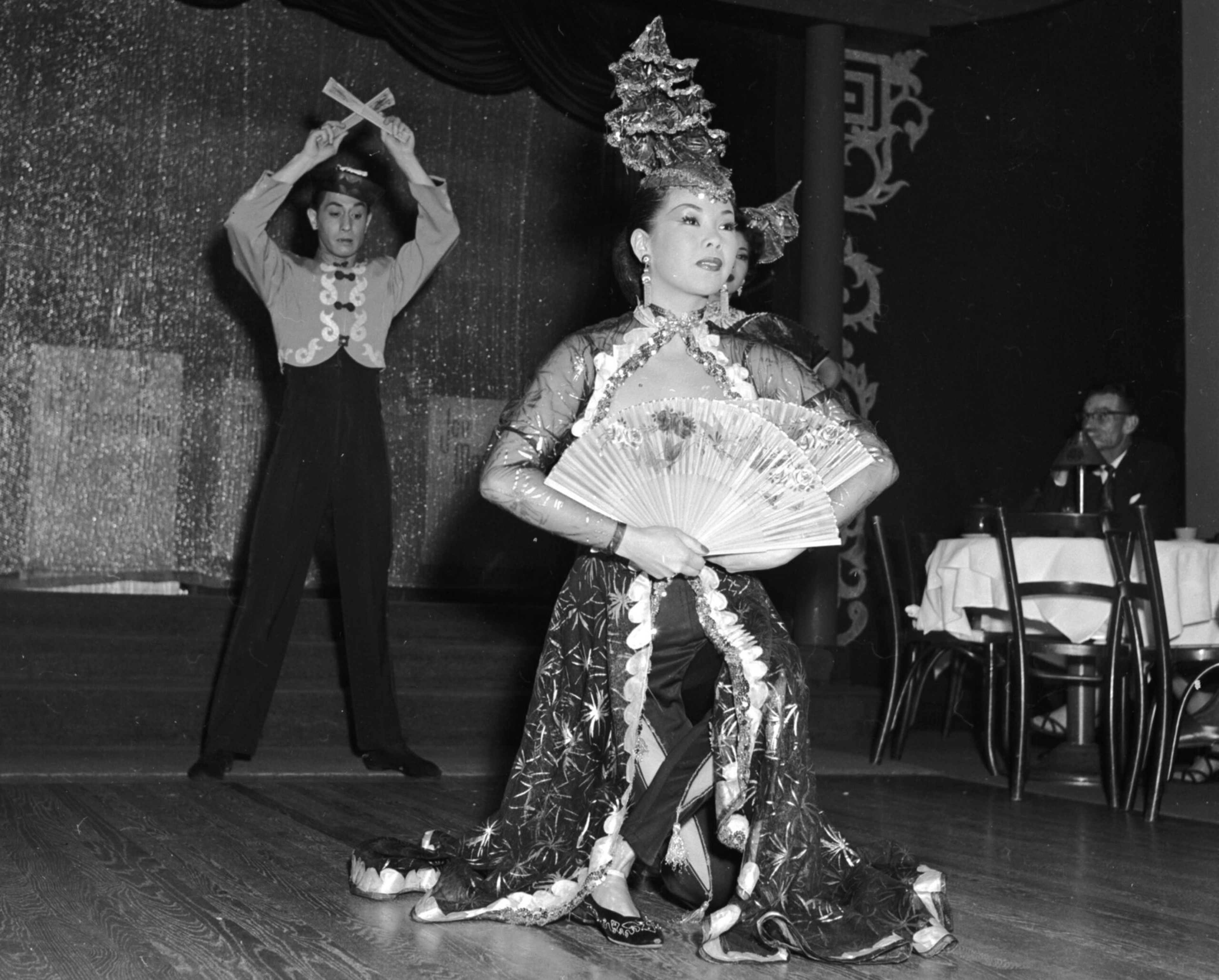 Chinese dancers perform on stage with elaborate costumes and a large decorate fan in a black-and-white photo. 