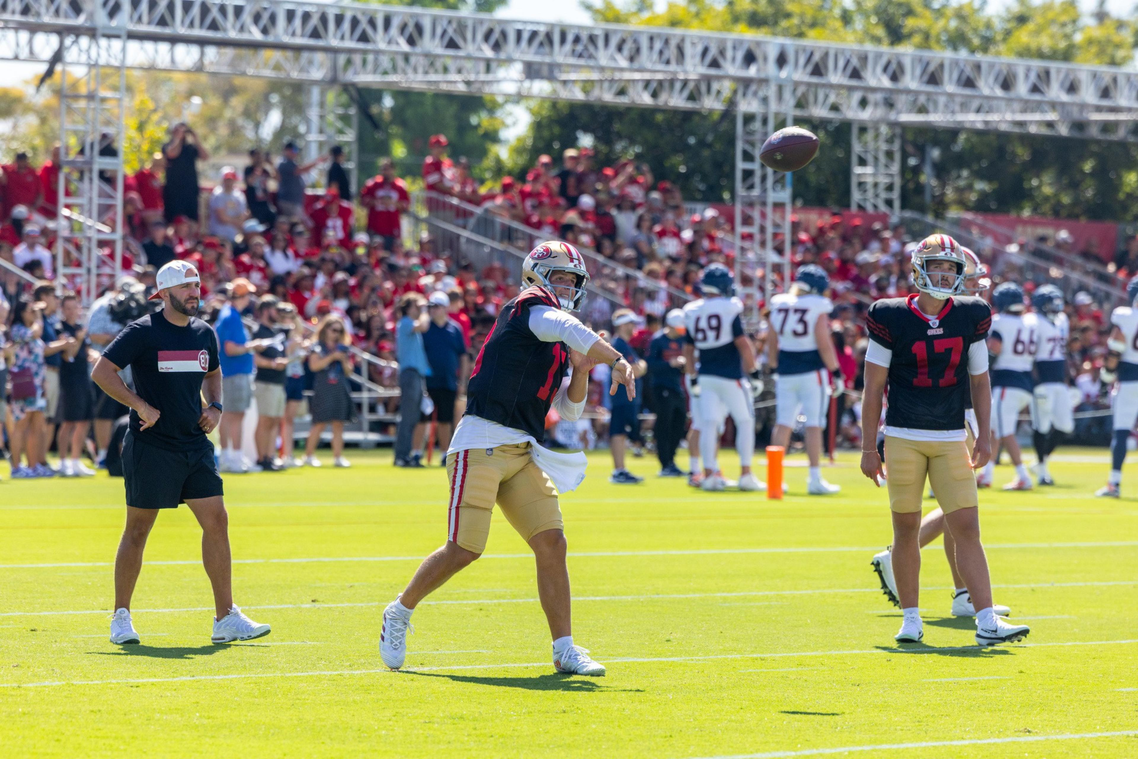 A football player in a black jersey and gold pants throws a football while teammates and a coach watch on a sunny field with spectators in the background.
