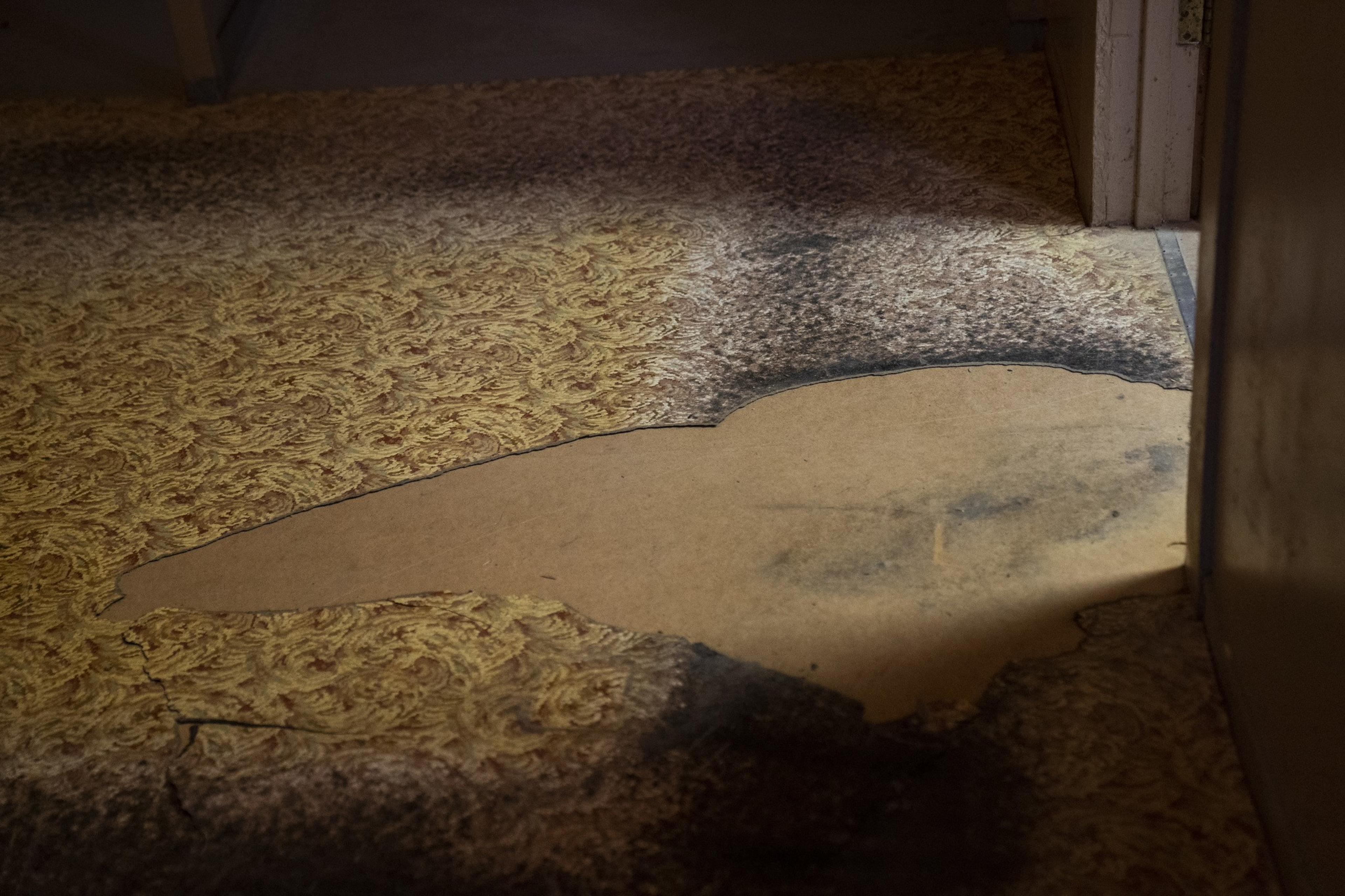 A worn floor shows a large, irregularly-shaped patch of exposed subfloor surrounded by aged, brownish, patterned linoleum.