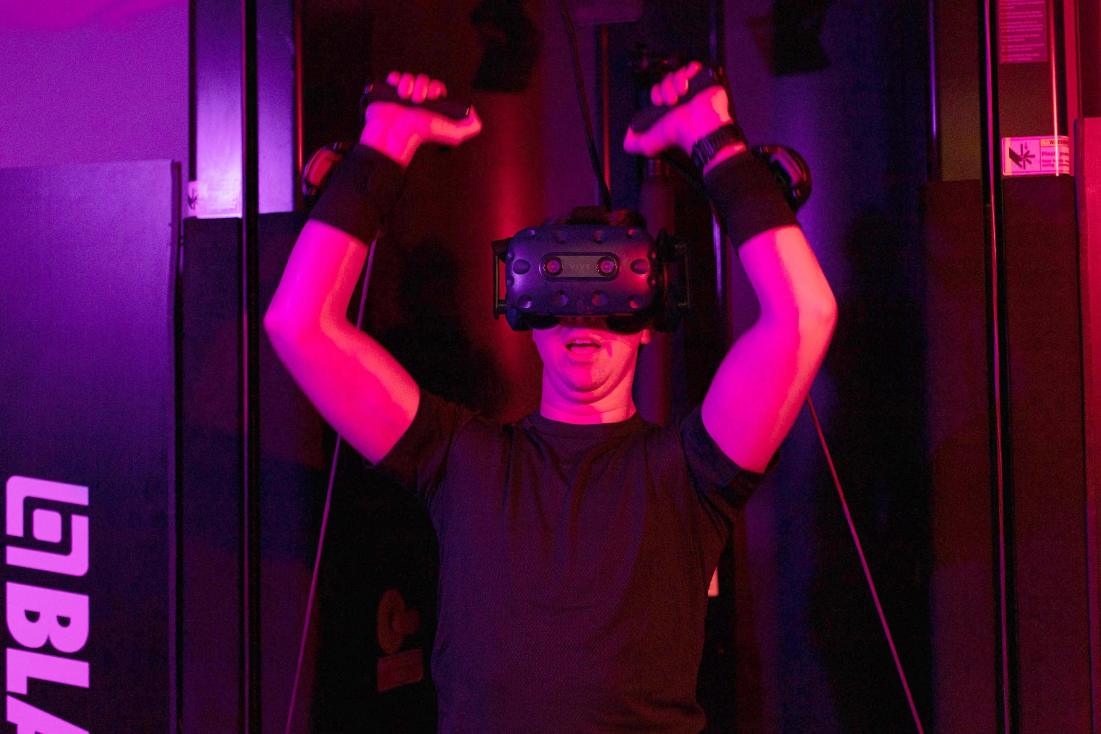 A person wearing a VR headset and gloves raises both hands, illuminated by purple and red lighting, immersed in a virtual reality experience.