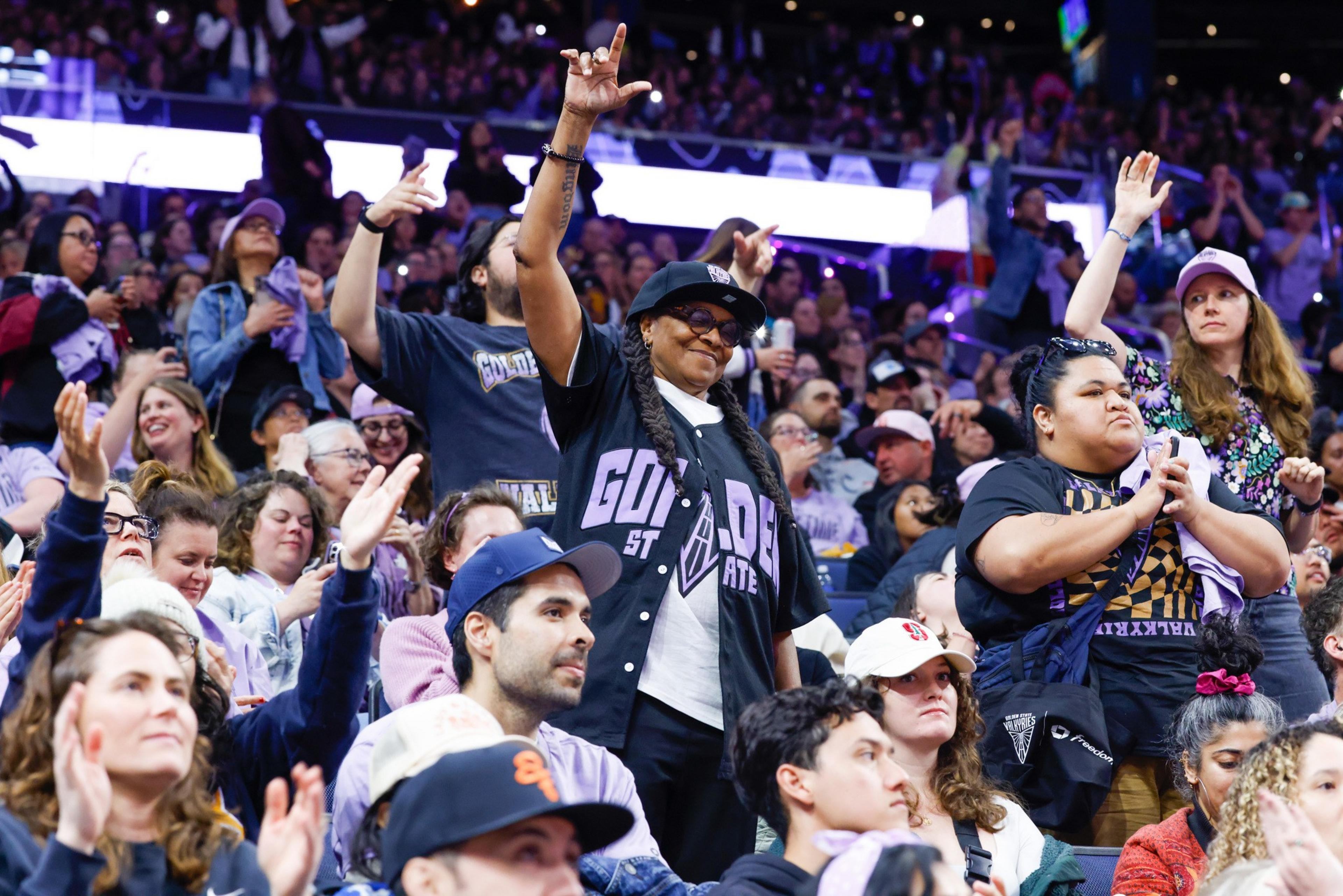 A lively crowd stands and cheers at a sports event, with fans in jerseys and hats. One person raises their hands, and others clap and take photos.