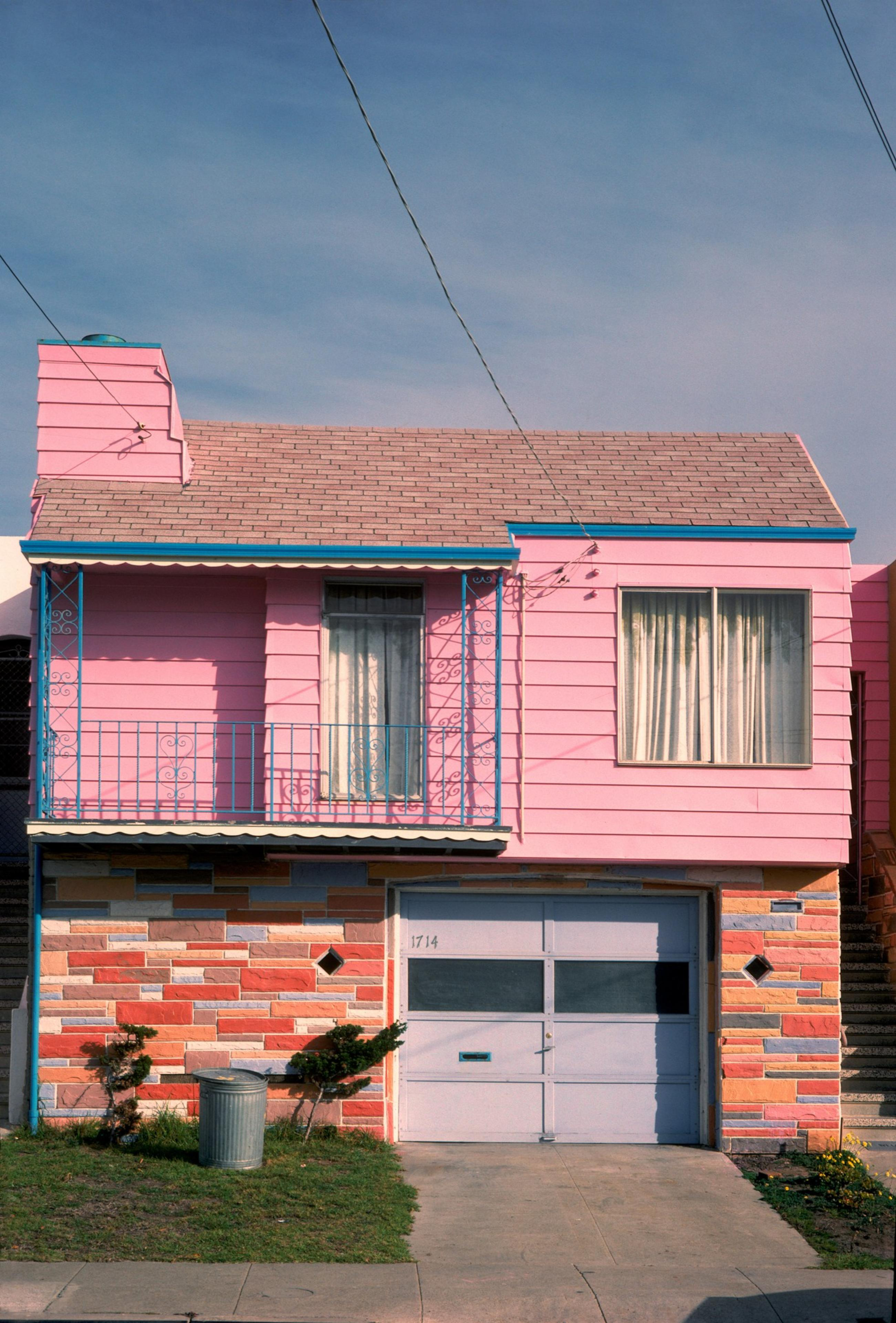 The image shows a two-story house painted pink with blue trim. It has a small balcony, multi-colored stone facade, garage, and a trash can on the front lawn.