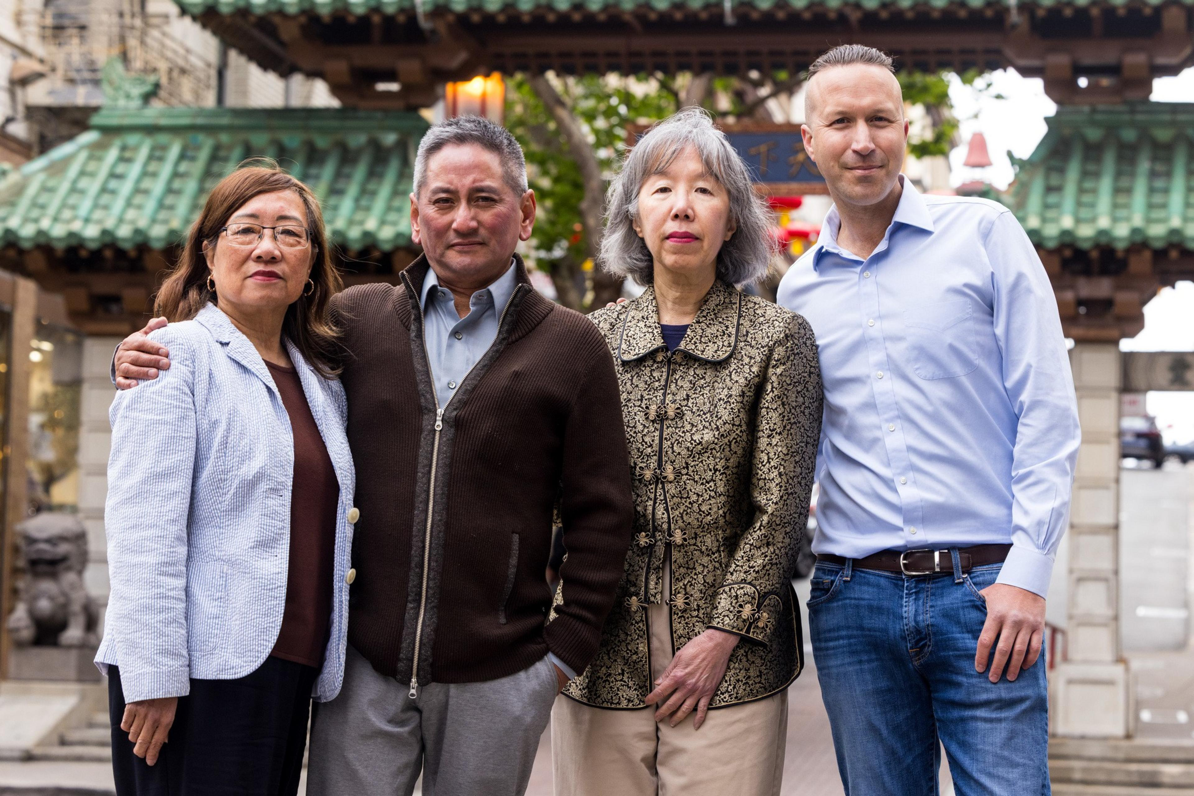 Four people stand together, smiling, with their arms around each other. They are outdoors, in front of a structure with green tiled roofs and stone details.