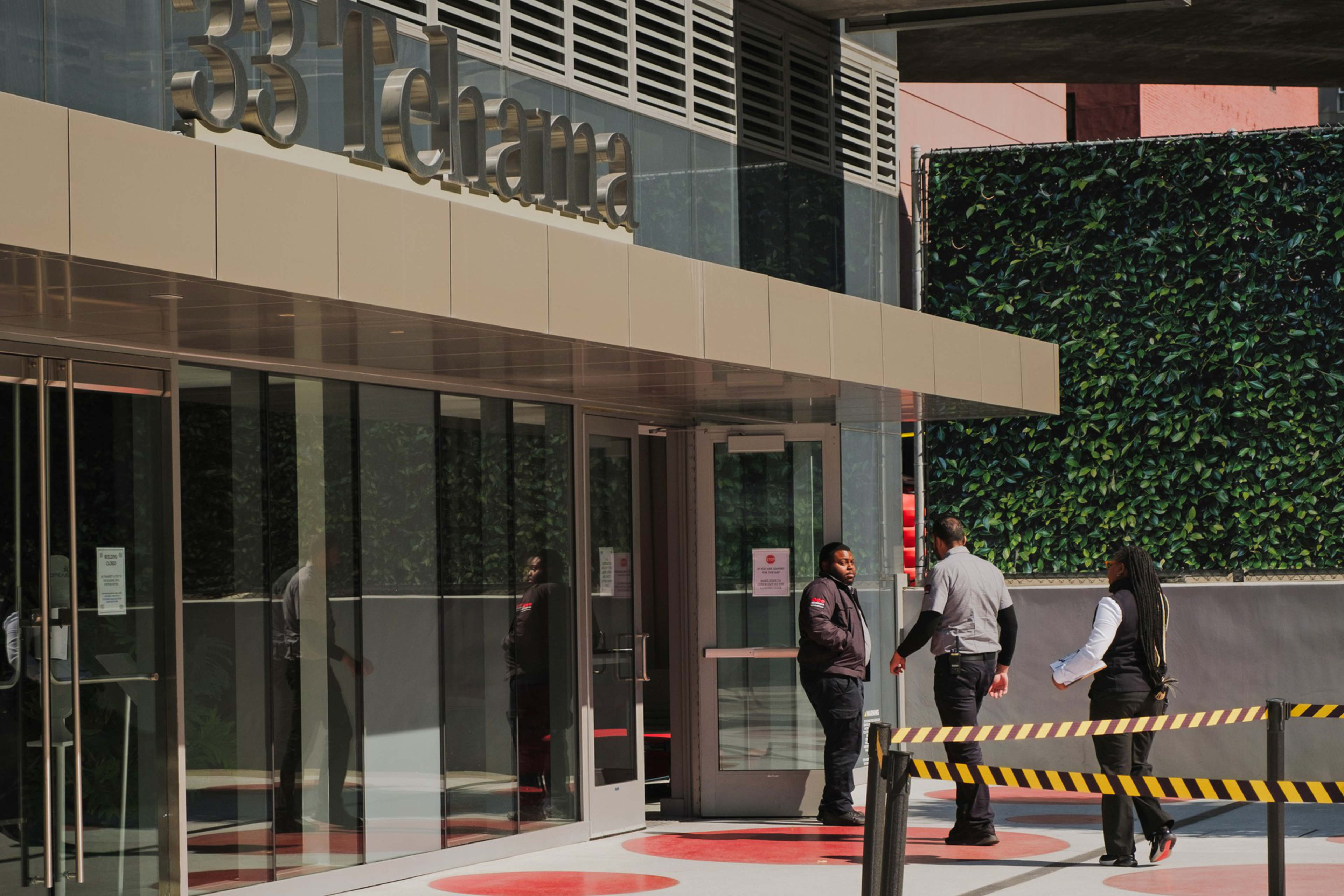The image shows a building entrance labeled "33 Tehama." Three people stand near the glass doors, with yellow-black caution tape forming a barrier nearby.
