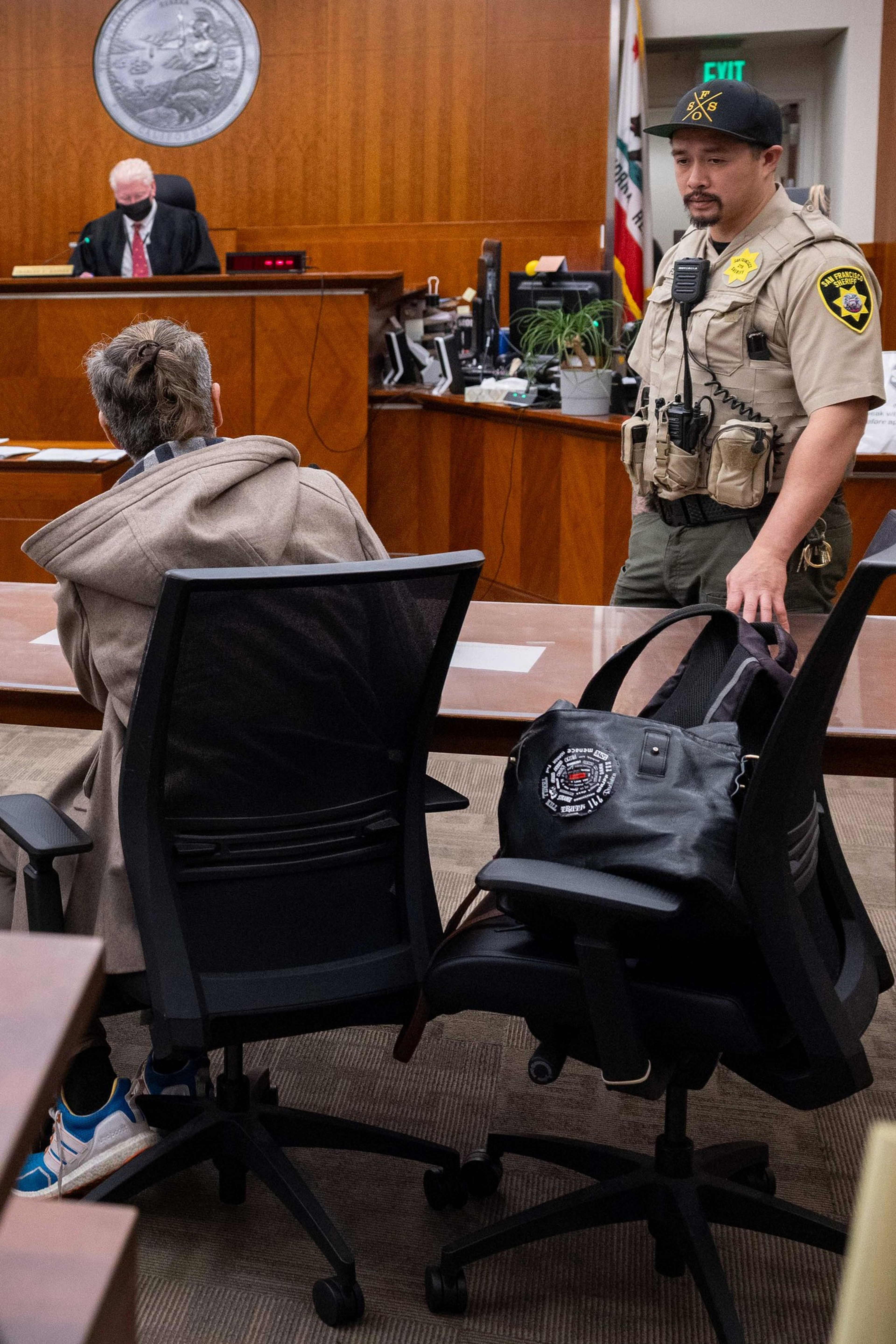 A man in a beige jacket and sneakers sits at a courtroom table, facing a judge, while a uniformed officer stands nearby with a black bag on a chair.