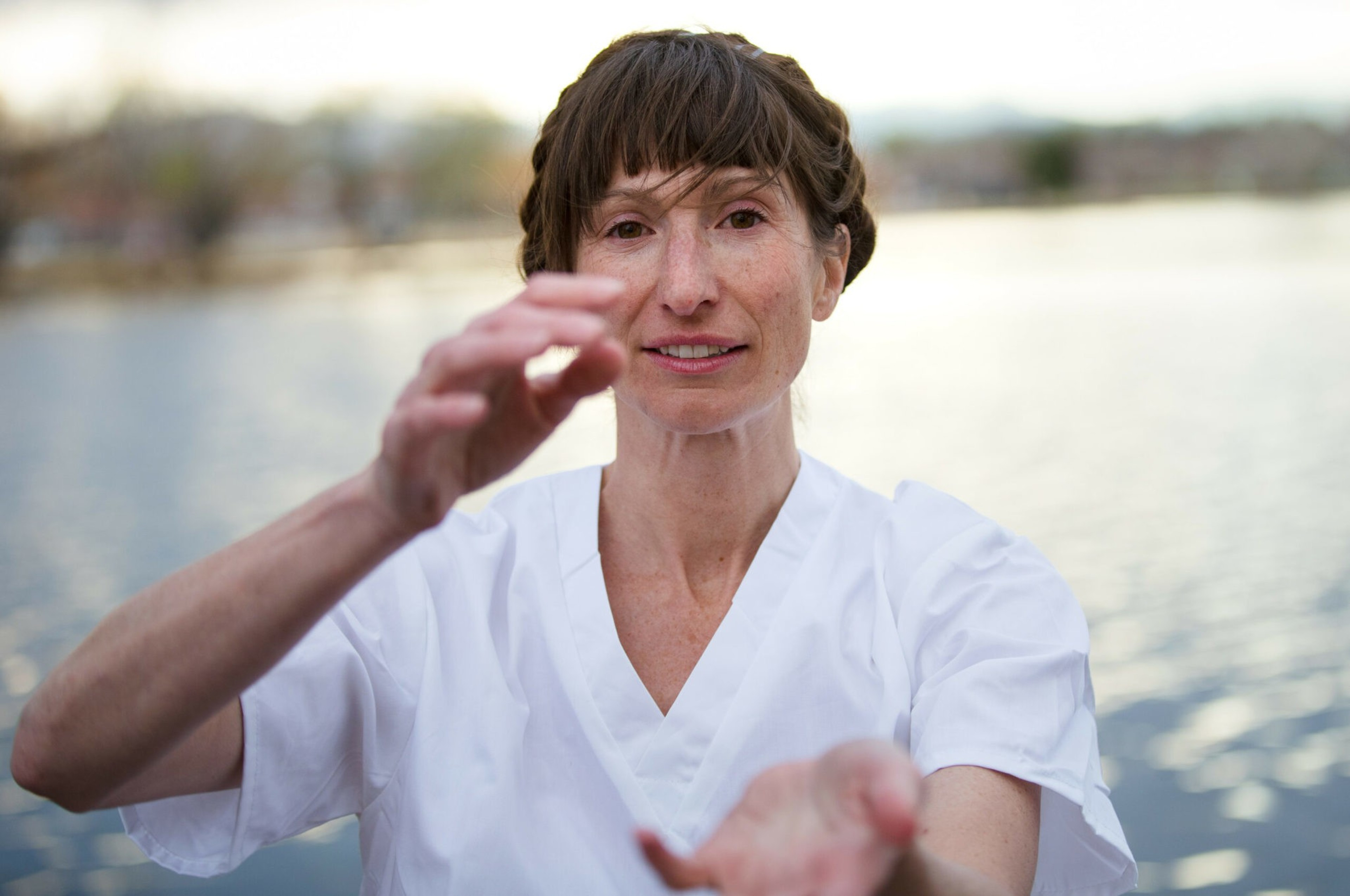 A woman in a white shirt stands near water, holding her hands up in a flowing, gentle pose, with a calm expression on her face.