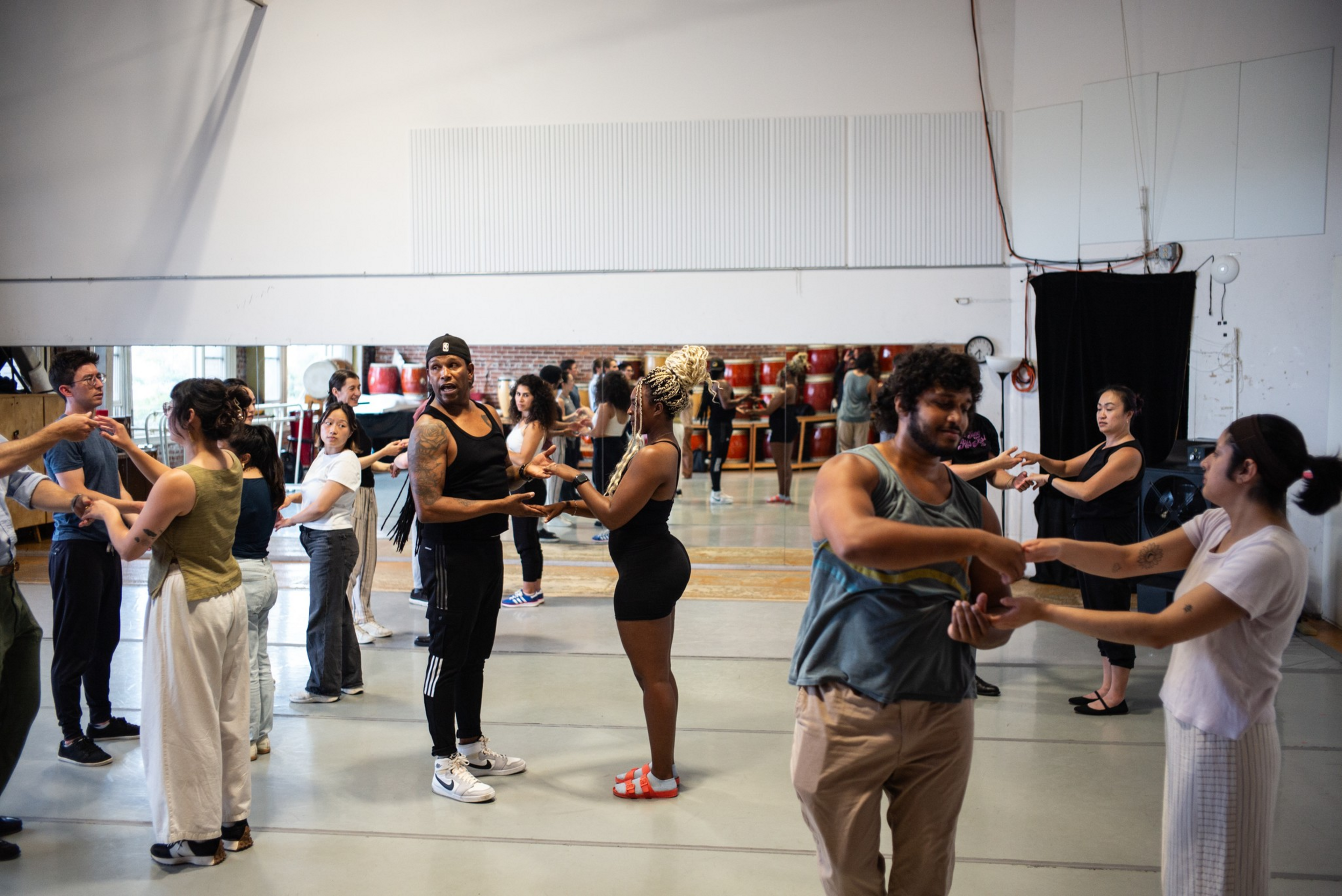 Several diverse pairs hold hands while standing in a dance studio with a large mirror, practicing partner dance steps under a bright, spacious setting.