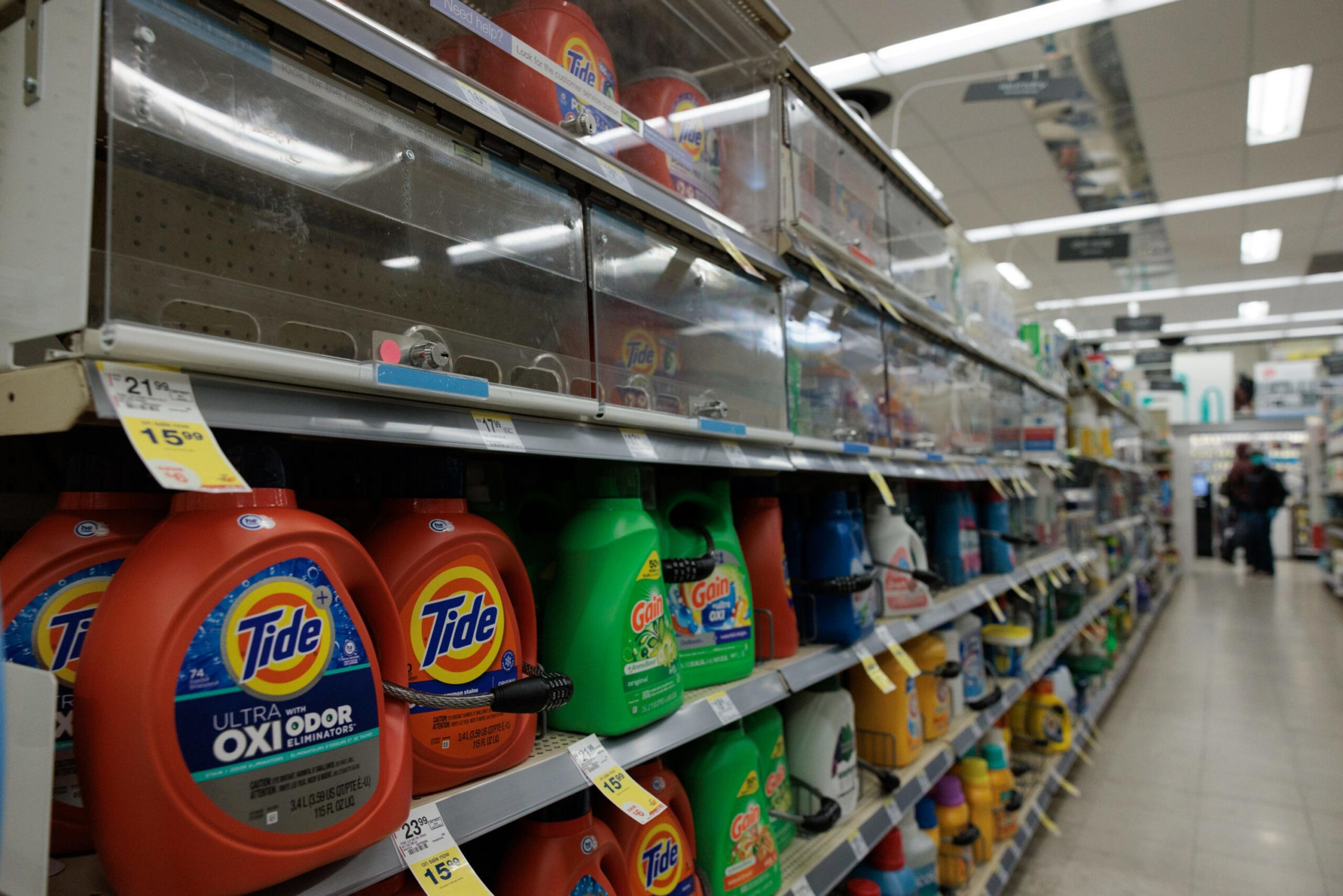 Laundry detergent bottles, including Tide and Gain, are displayed on store shelves with price tags and security locks attached.