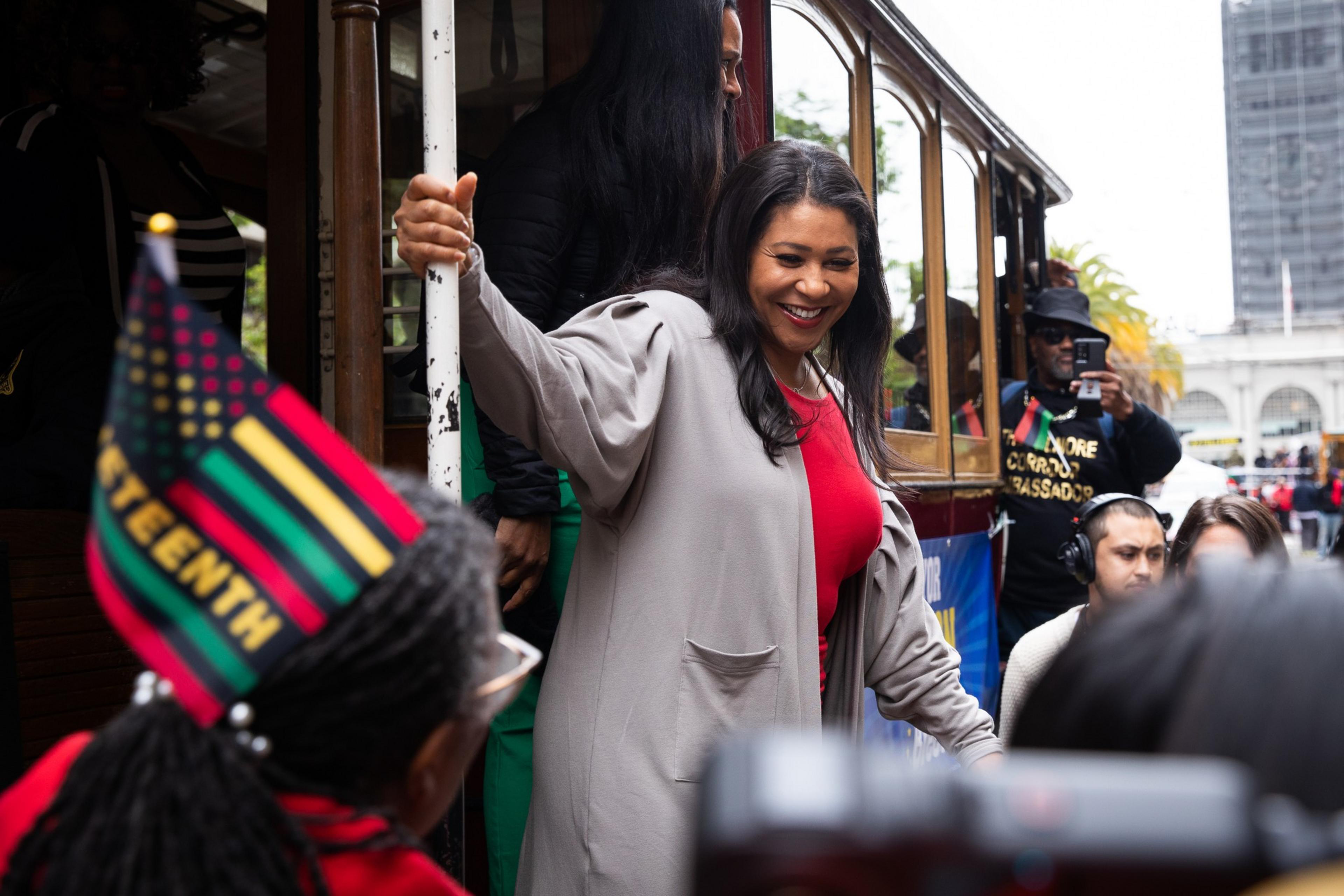 A smiling woman in a gray coat and red top steps off a wooden trolley, while a person nearby wears a flag-themed Juneteenth hat.