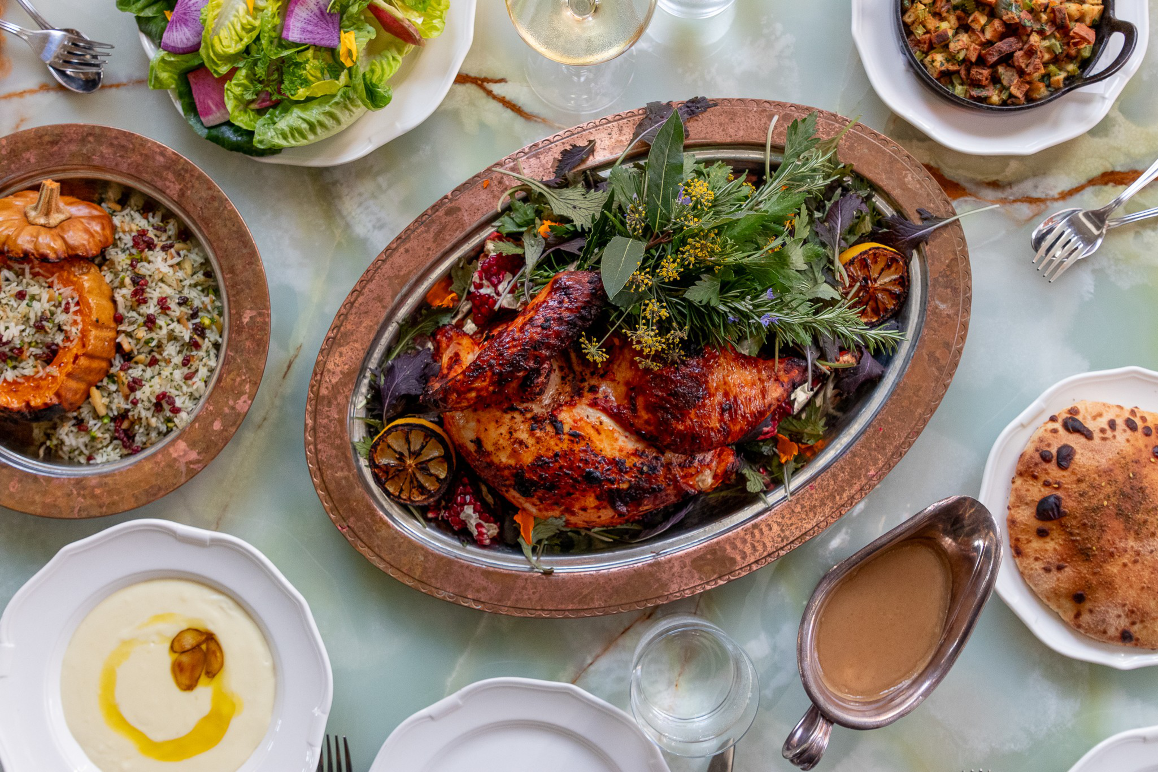 A roasted whole chicken garnished with herbs and lemon sits on a platter, surrounded by bread, salad, grains, gravy, and creamy sauce on a marble table.