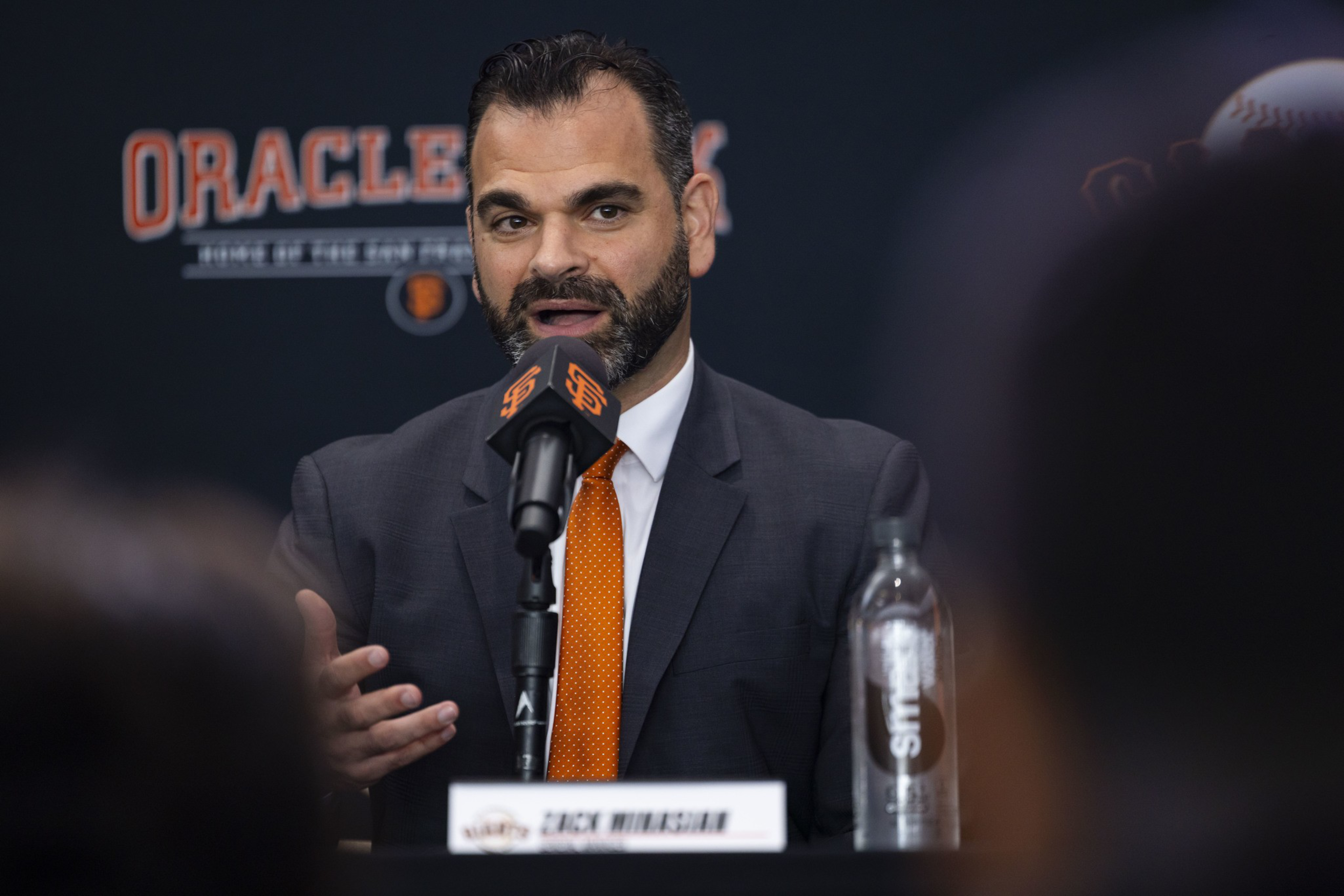 A man in a dark suit and orange tie speaks at a microphone with San Francisco Giants logos and a water bottle nearby.